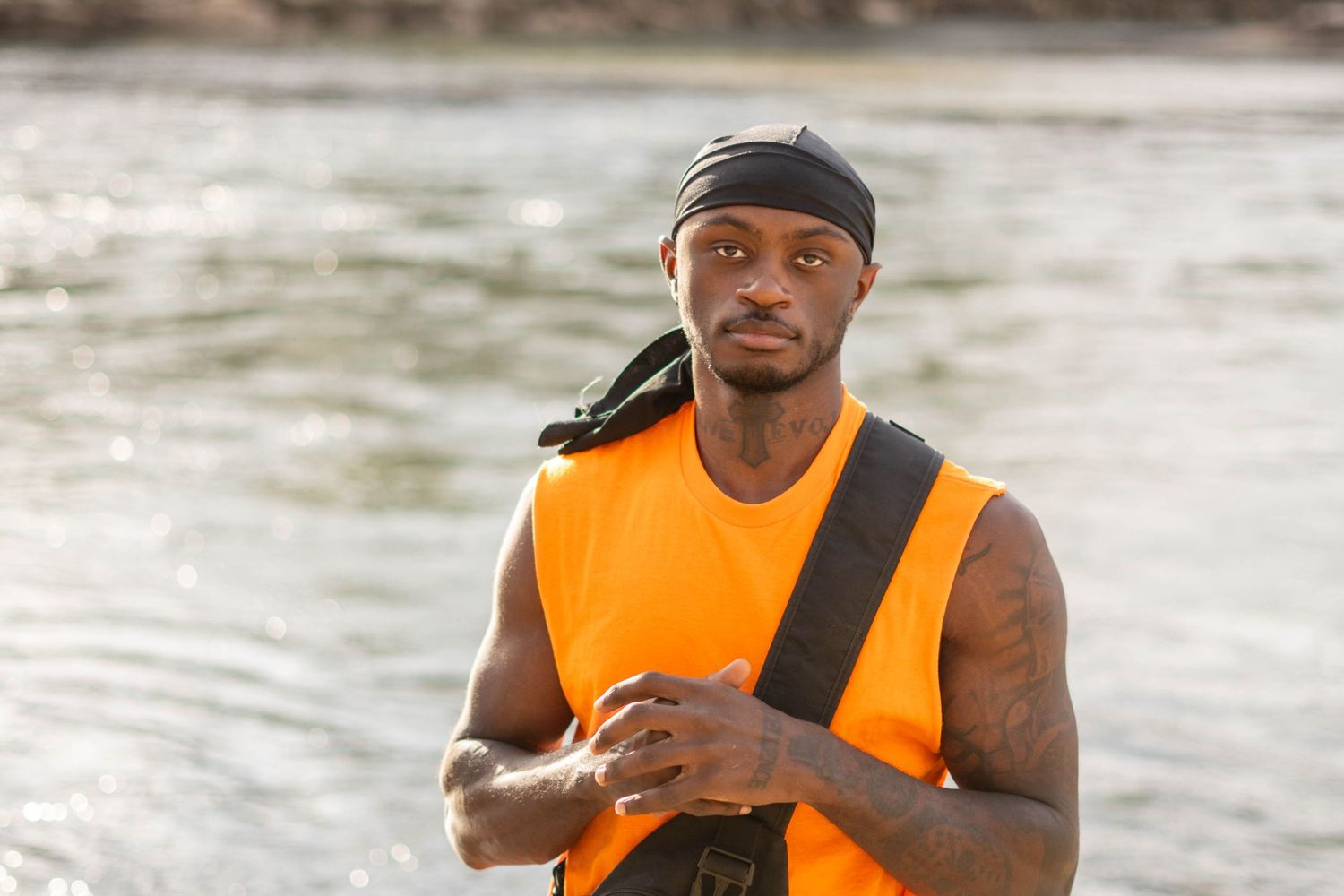Athletic person in orange safety vest and durag standing by water on sunny day.