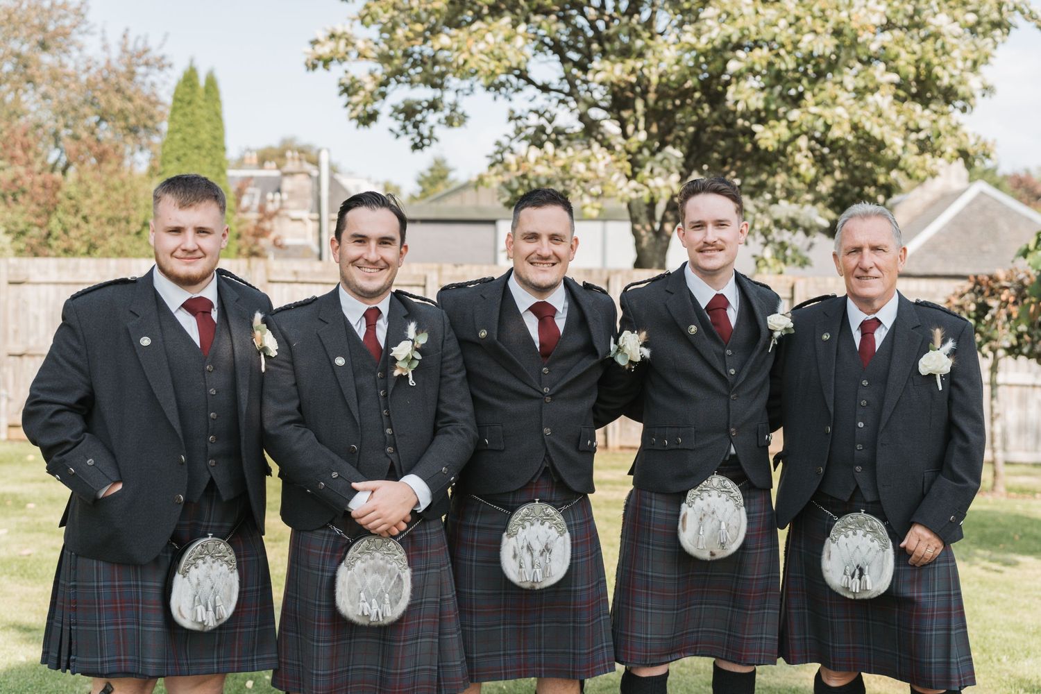 Five groomsmen in traditional Scottish kilts and black jackets pose together outdoors on a wedding day.