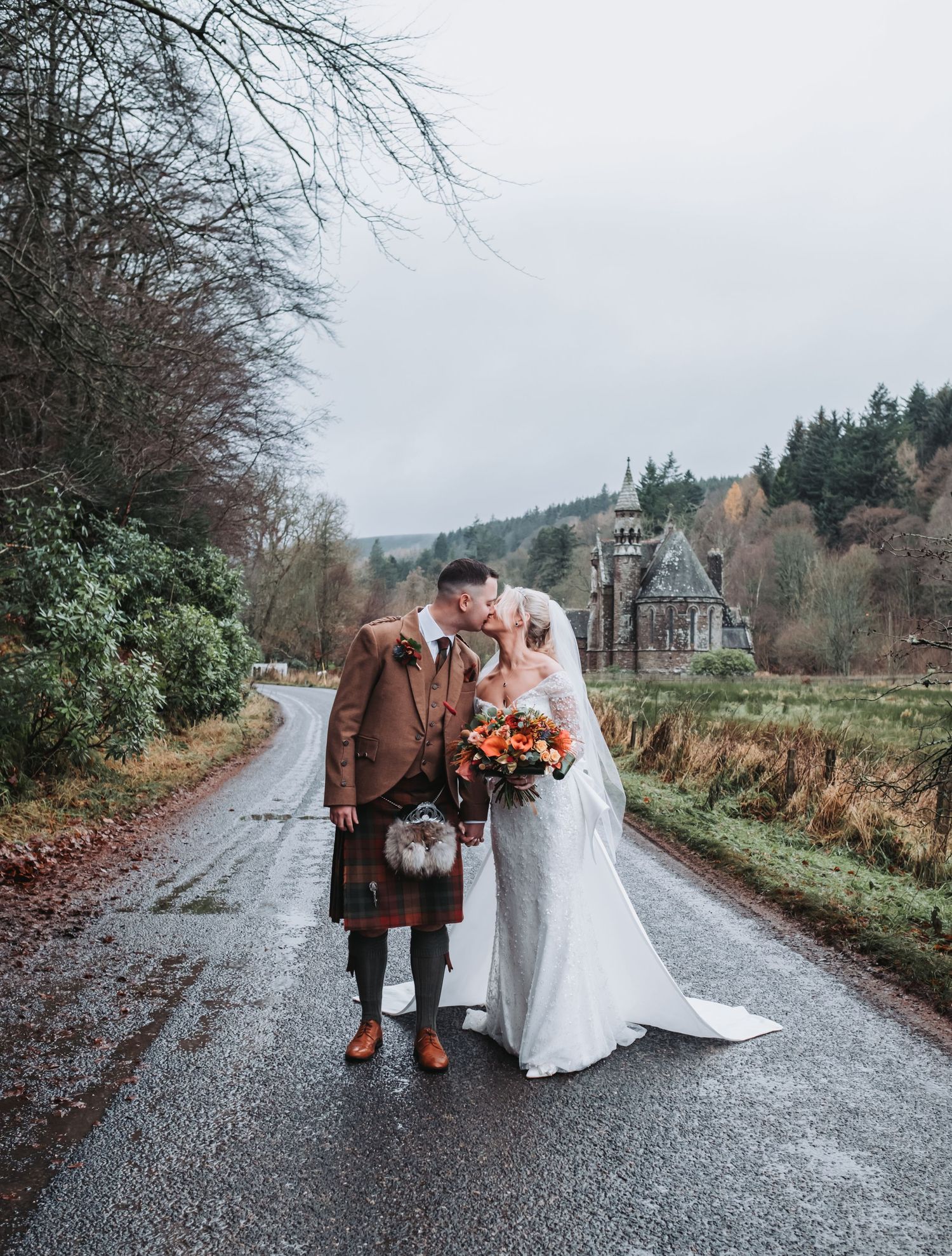 A bride and groom kissing on a rainy country road in Scotland with a castle visible in the background.