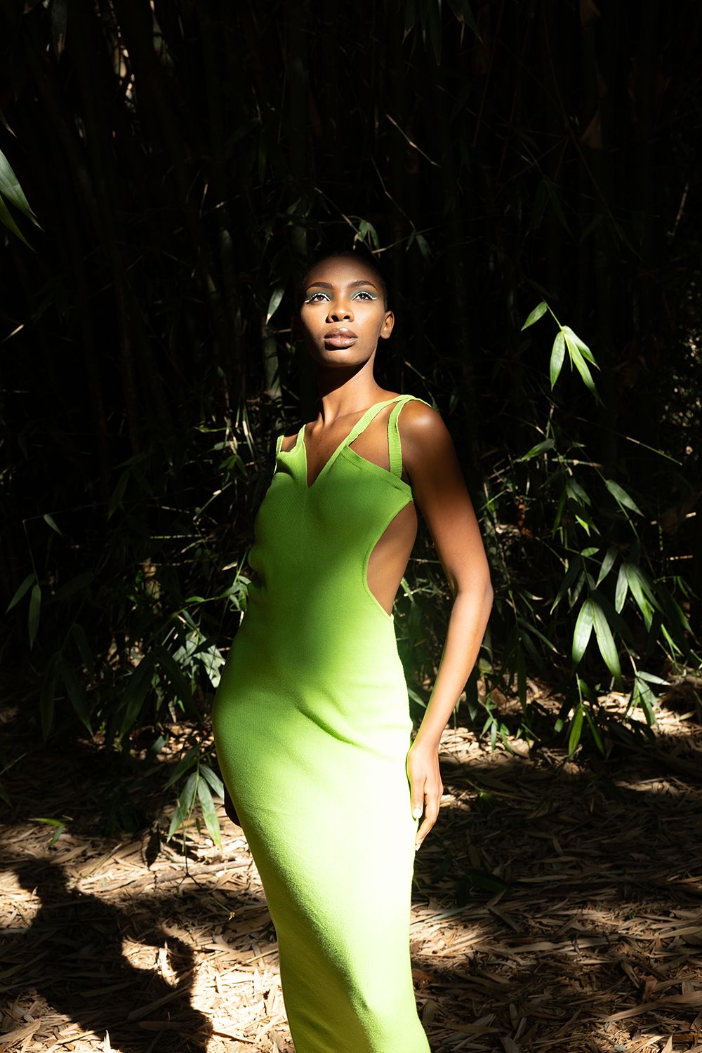 Fashion model poses in form-fitting lime green dress against dark foliage background in dramatic lighting.