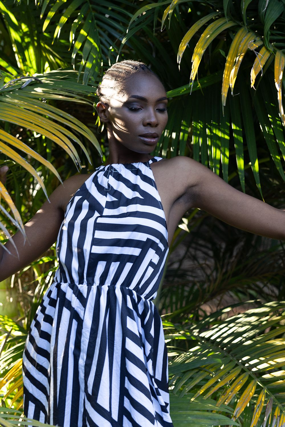 Elegant model wearing black and white striped halter dress poses against bright green and yellow tropical palm leaves. Model looks down/away
