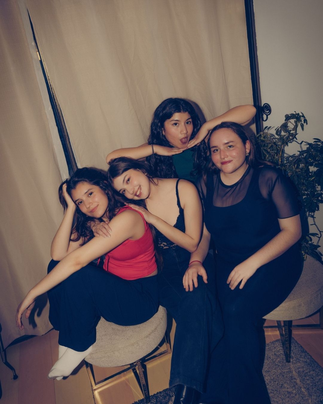 Four young women posing playfully together in a casual indoor setting with curtains and plants.