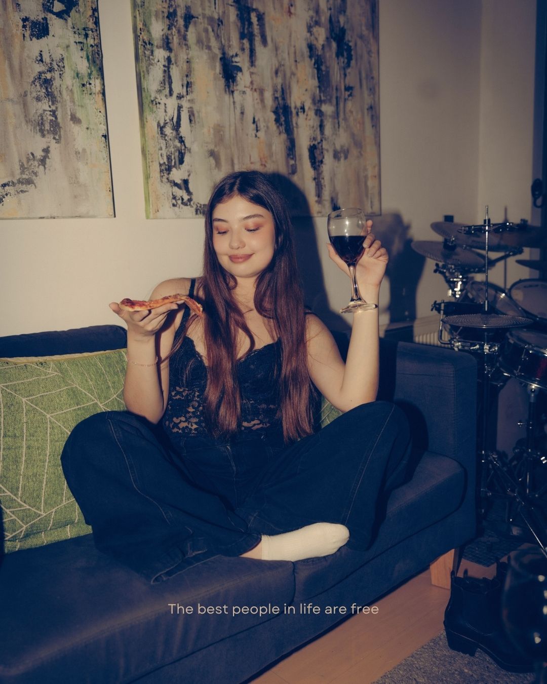 Young woman sitting cross-legged on a couch holding pizza and wine glass in a cozy room.