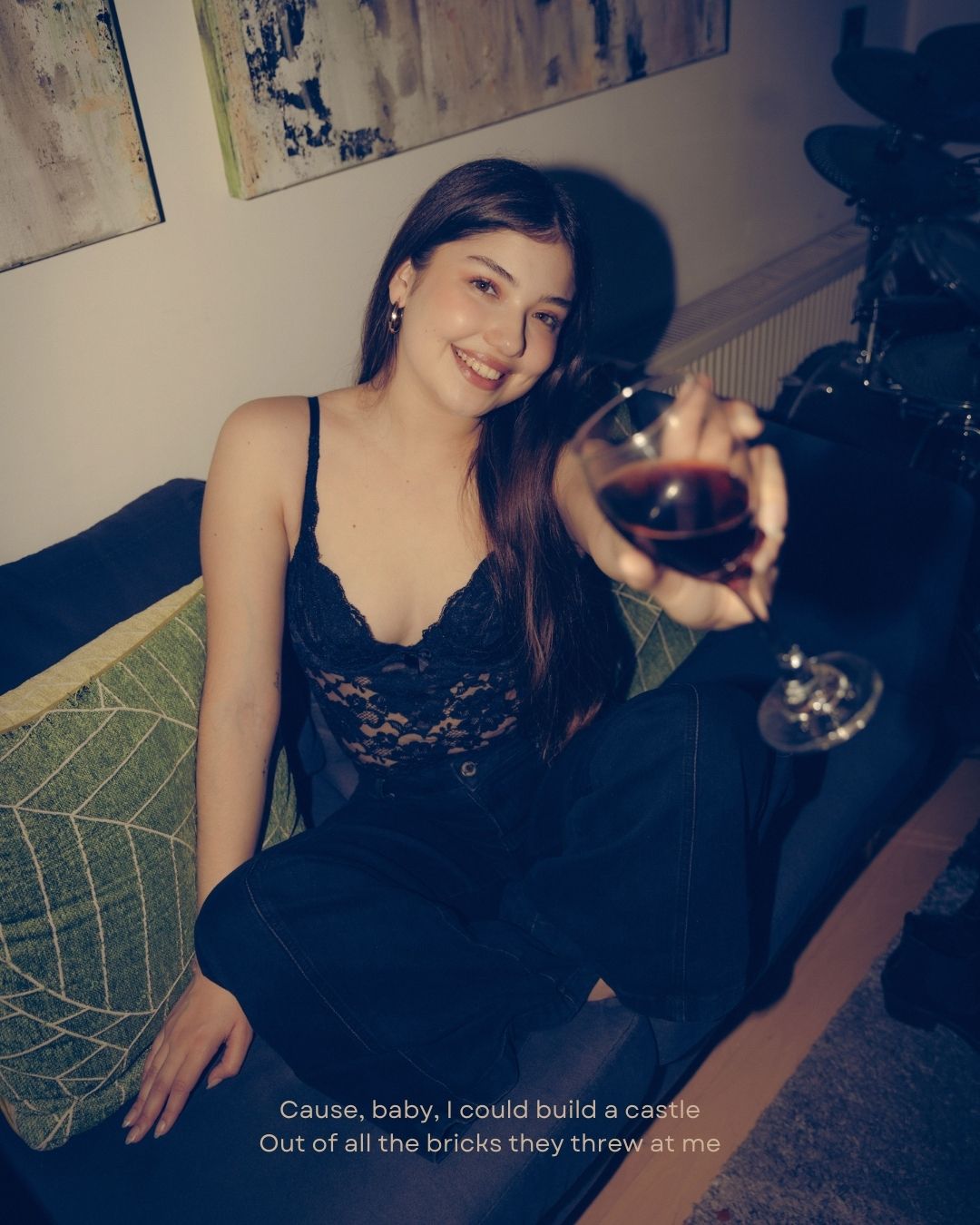 Young woman in black lace top smiling while holding a glass of red wine at a cozy indoor party.
