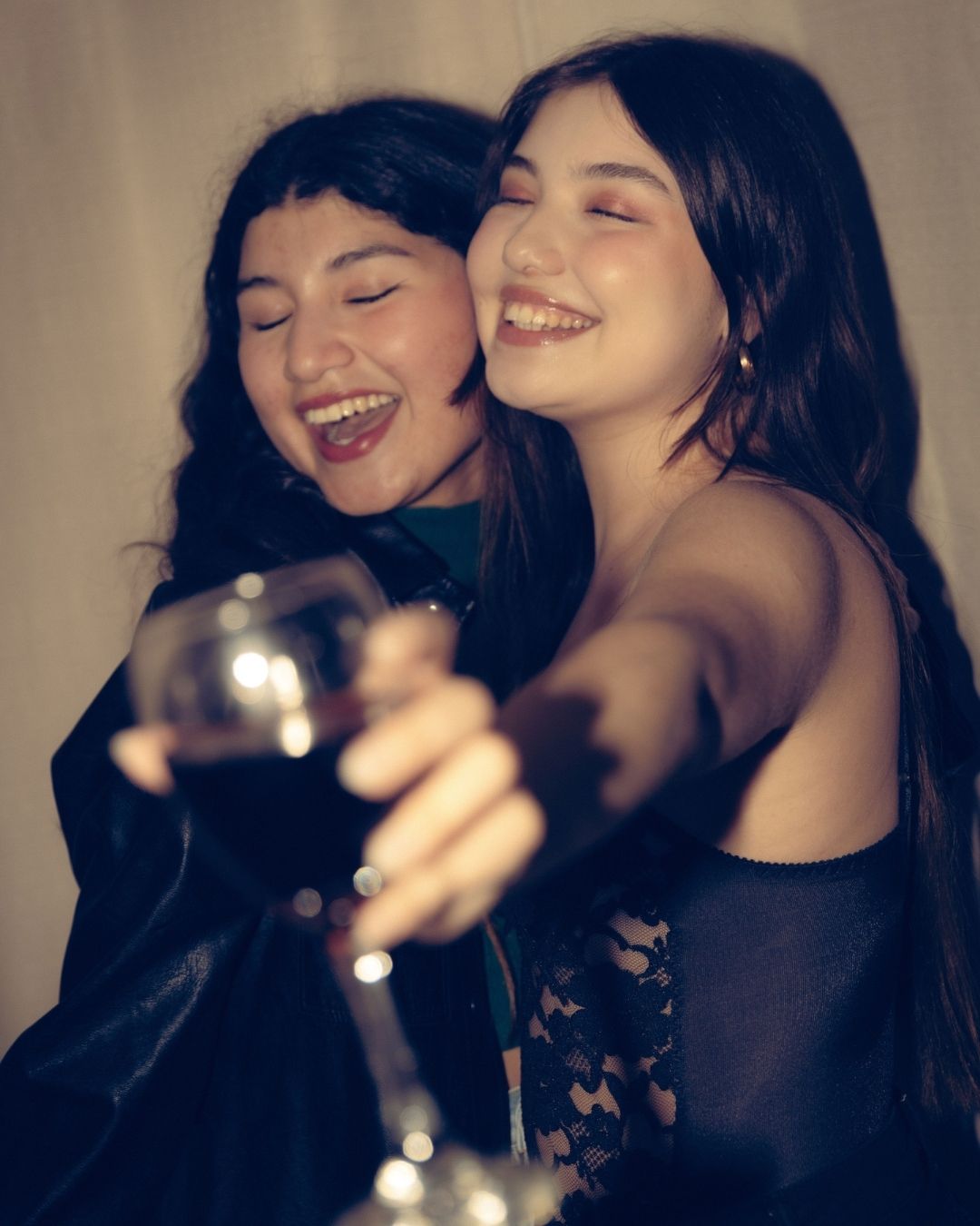 Two young women laughing joyfully while toasting with a glass of red wine at a party.