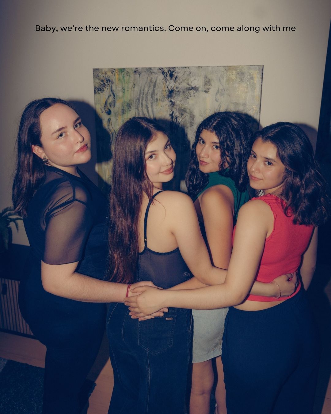 Four young women posing together in stylish outfits at an indoor gathering with artwork on the wall.
