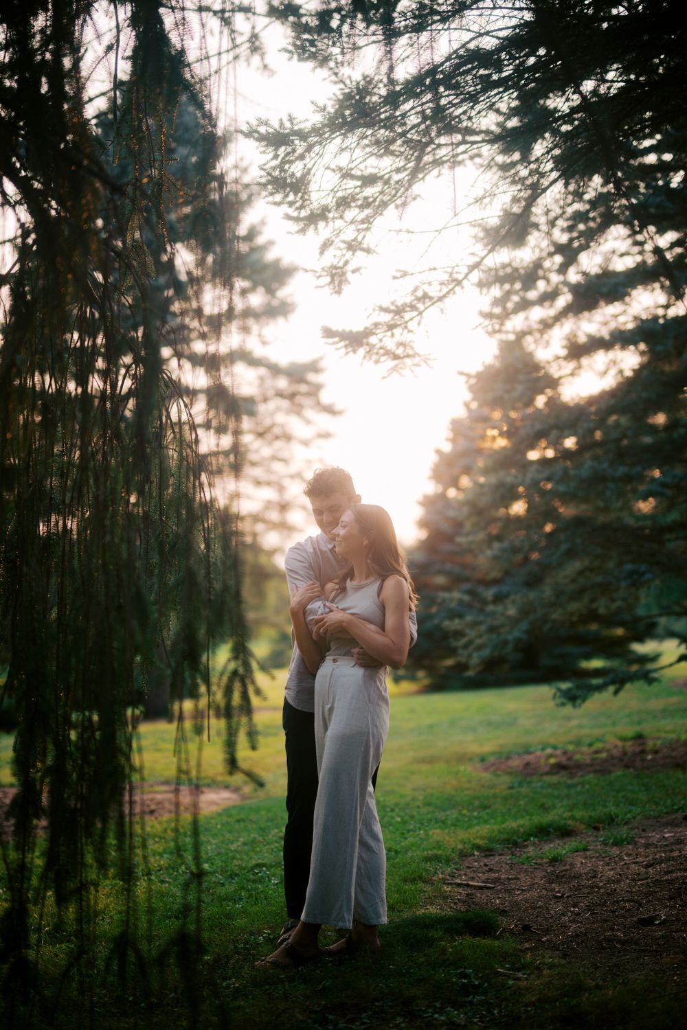 A silhouetted figure stands holding a baby against a misty sunset backdrop among pine trees in a peaceful garden setting.
