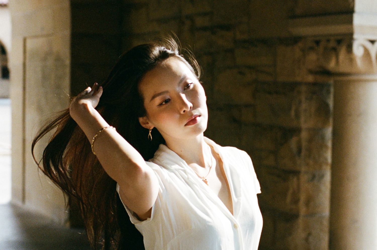 A young woman in a white blouse poses naturally by a stone wall while gently touching her flowing dark hair.