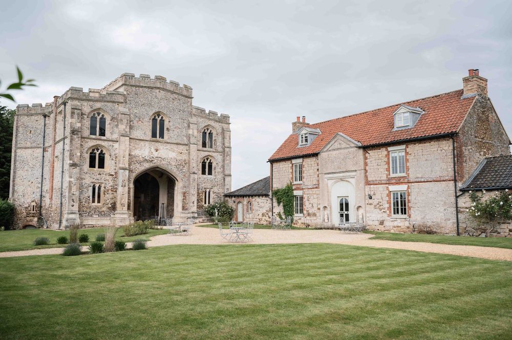 Historic stone castle gatehouse with Norman arches and tower stands beside a rustic brick house with a manicured lawn.