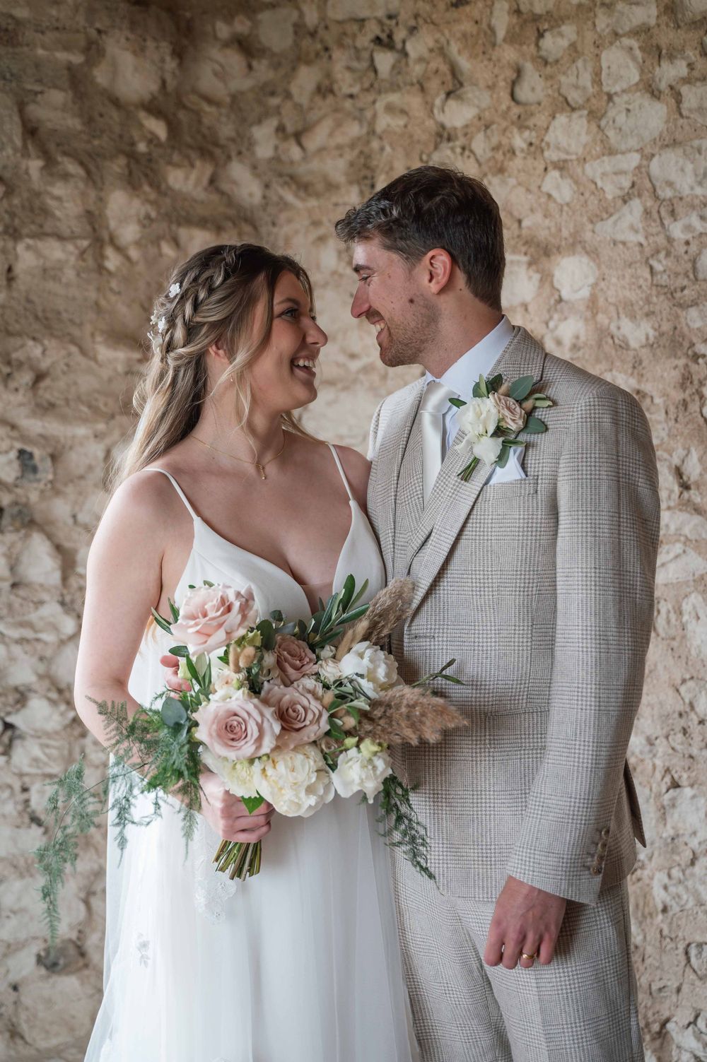 A bride and groom share an intimate moment against a rustic stone wall while holding a soft pink and white floral bouquet.