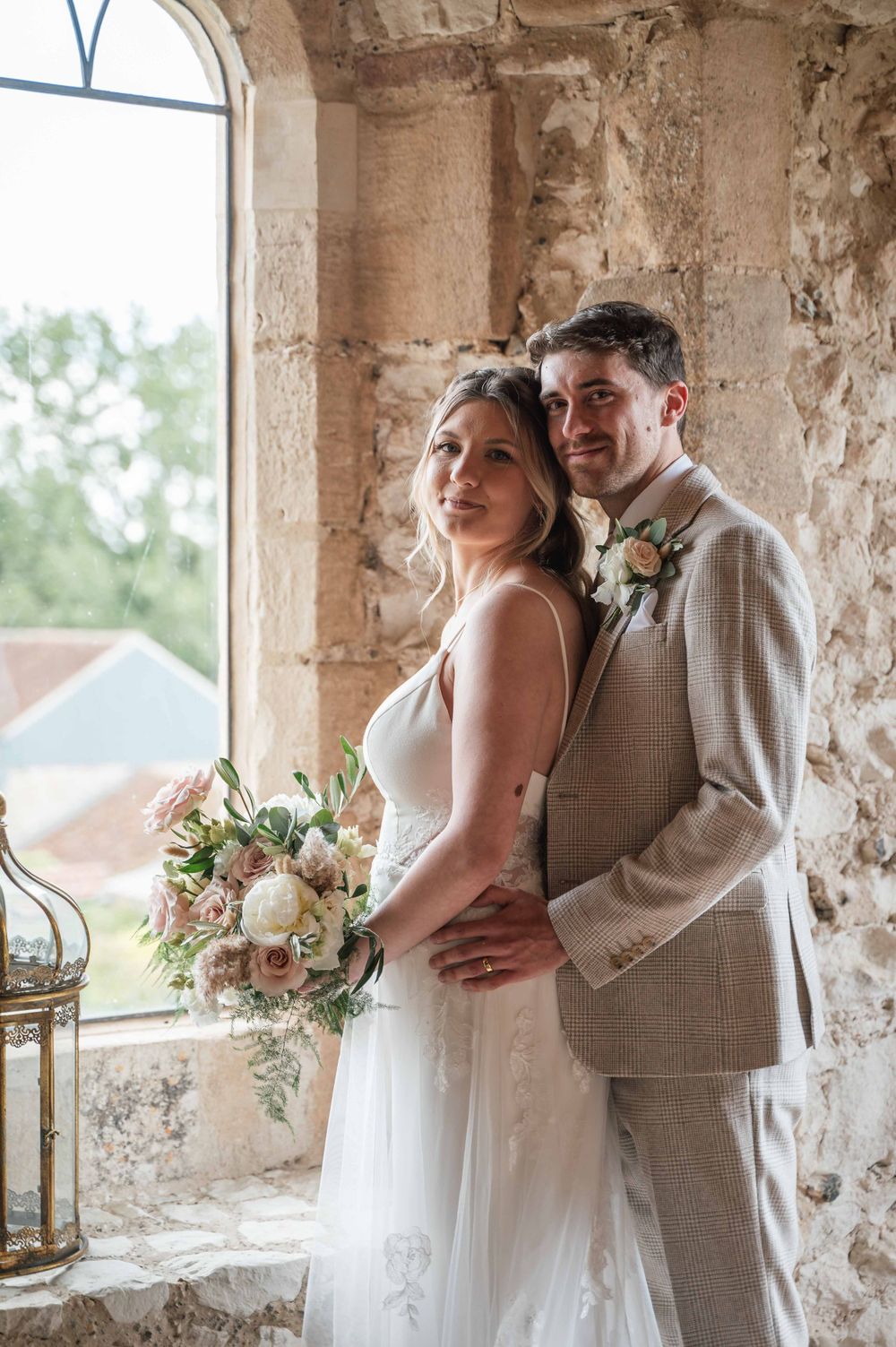 A romantic wedding couple embraces in an elegant stone archway with weathered walls and a soft bouquet of pink flowers.