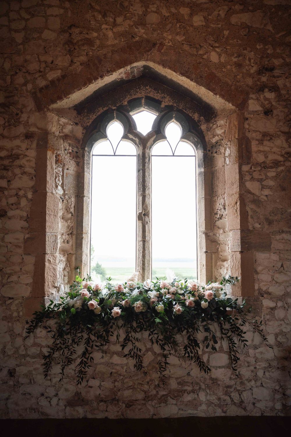 Gothic stone window arch adorned with delicate pink and white roses and greenery in a medieval church setting.