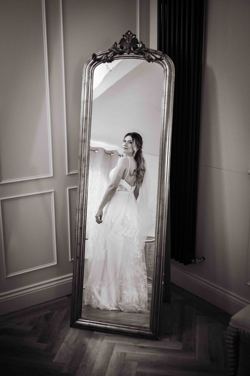 A vintage ornate floor mirror in a room with wood flooring reflects an elegant white wedding gown.