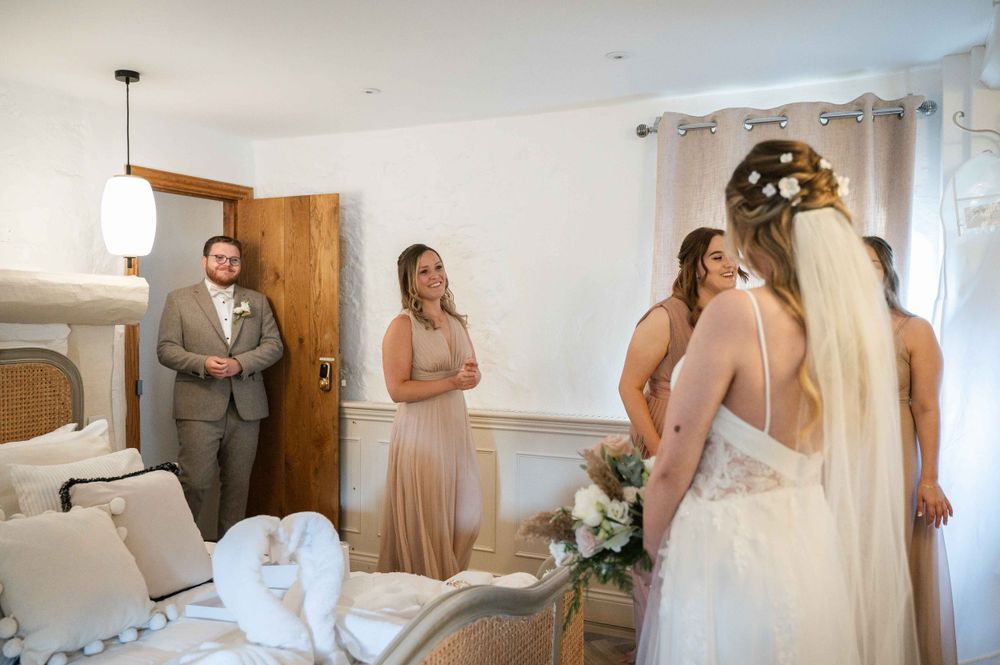 A group gathers in a bright room with bridesmaids in champagne dresses and a bride in white preparing for a wedding.