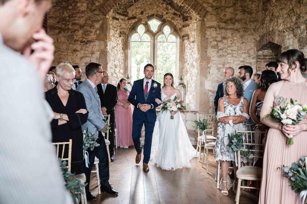 A wedding ceremony taking place inside a rustic stone chapel with gothic arched windows and elegant floral decorations.