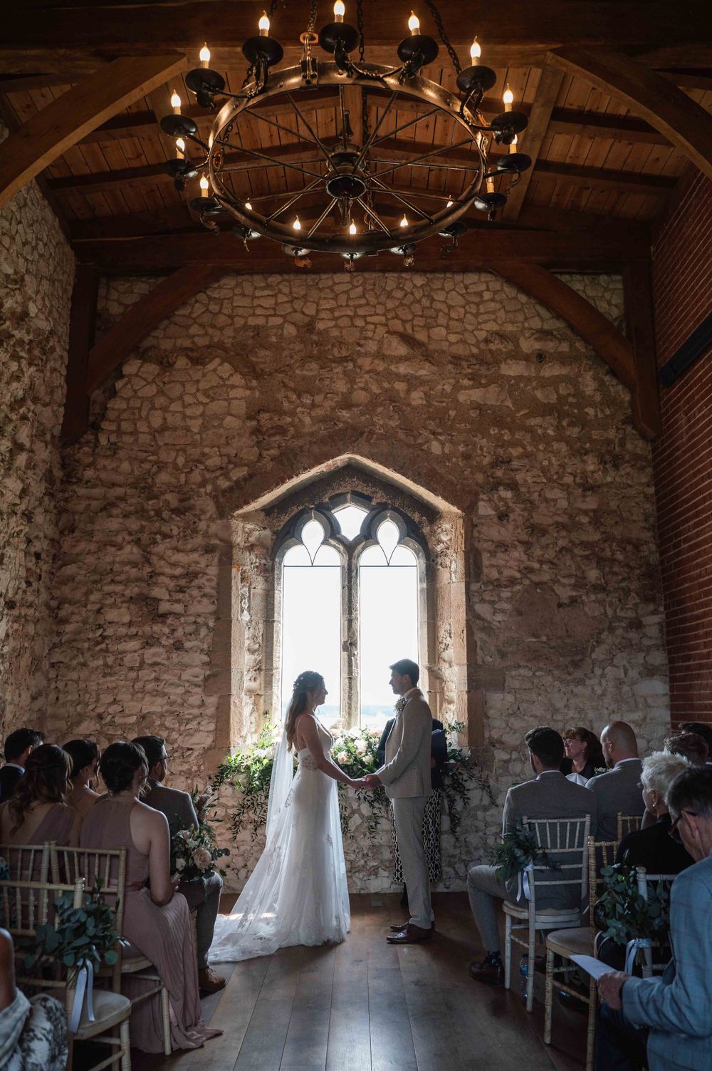 A romantic wedding ceremony in a rustic stone chapel with gothic windows and a circular chandelier overhead.