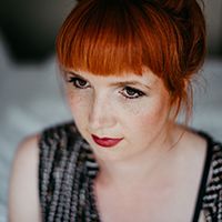 Frau mit roten Haaren und Pony schaut nachdenklich zur Seite. Sie trägt ein gemustertes Oberteil und roten Lippenstift.