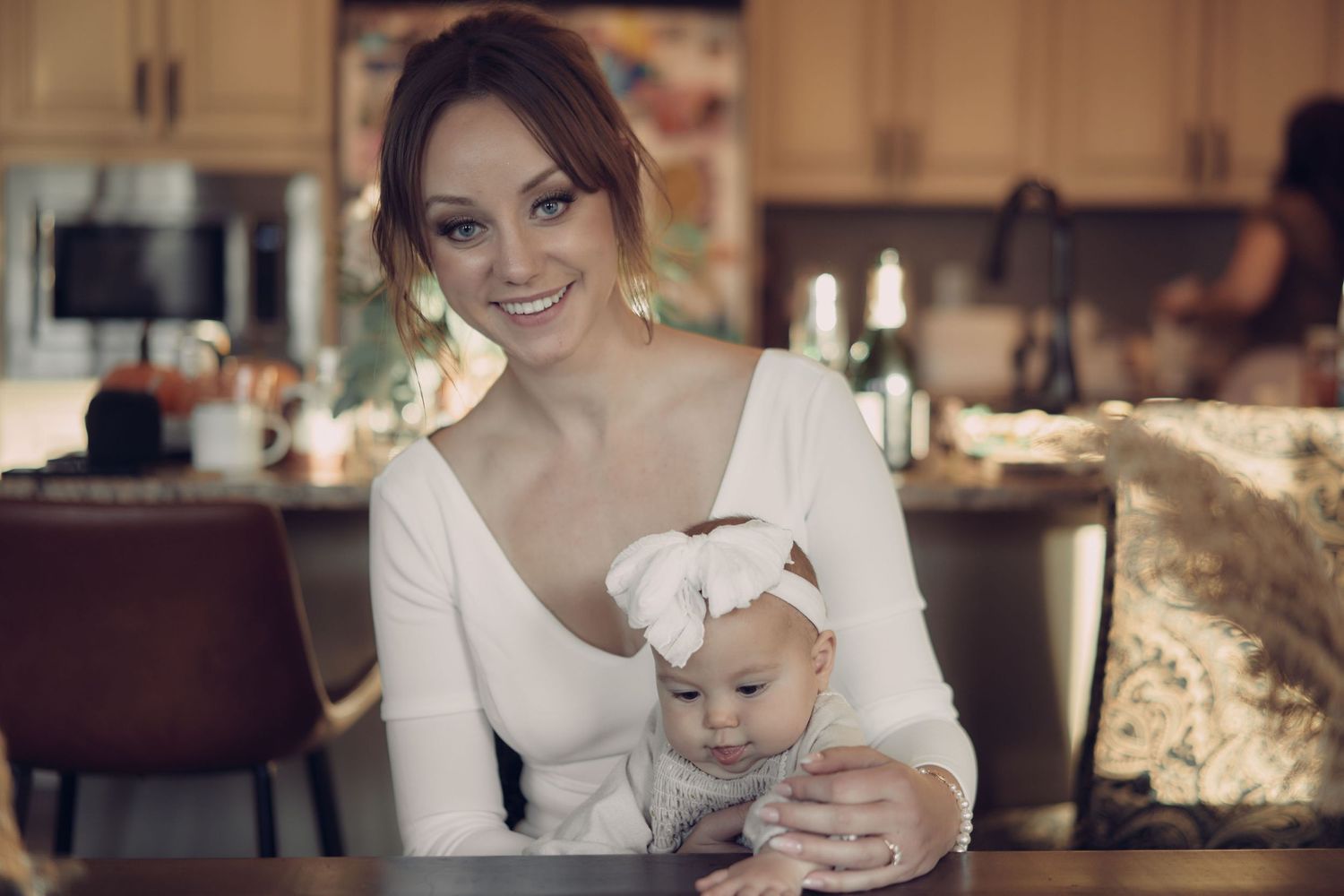 A bride in a white dress smiles warmly while holding a baby with a bow headband, sharing a tender moment in a cozy, softly lit kitchen.