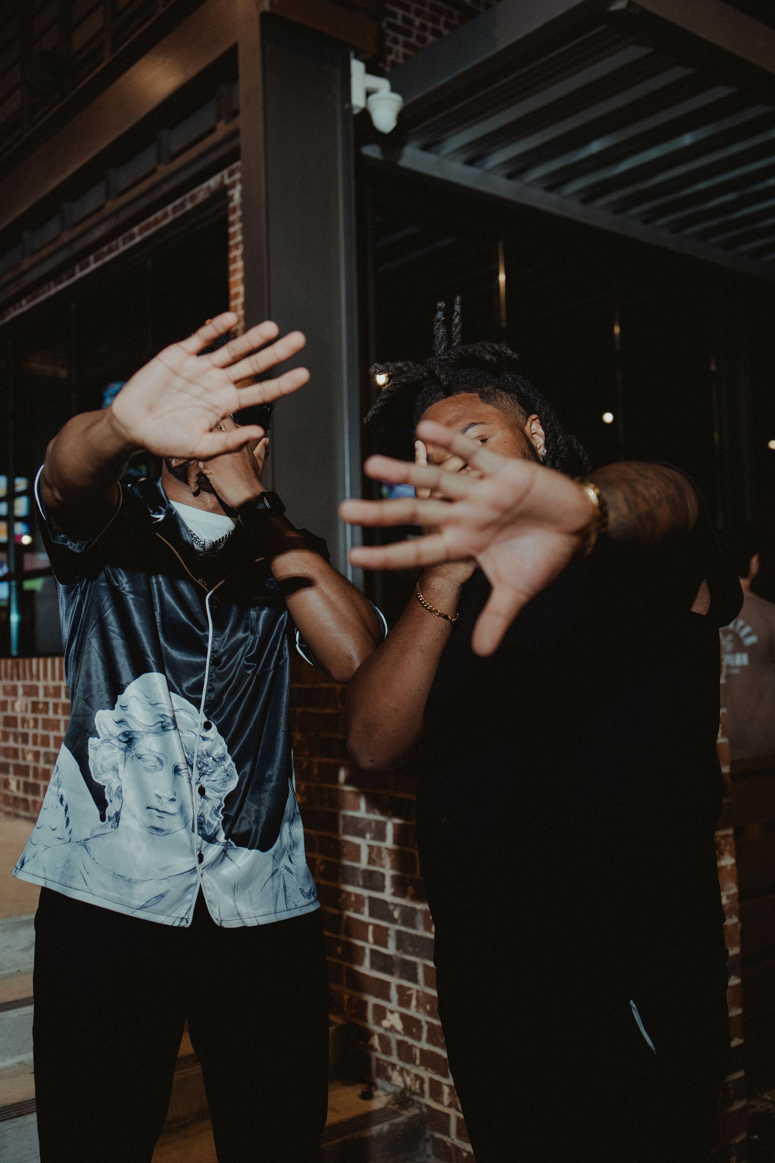 Two people playfully blocking their faces with their hands while standing against a brick wall at night.