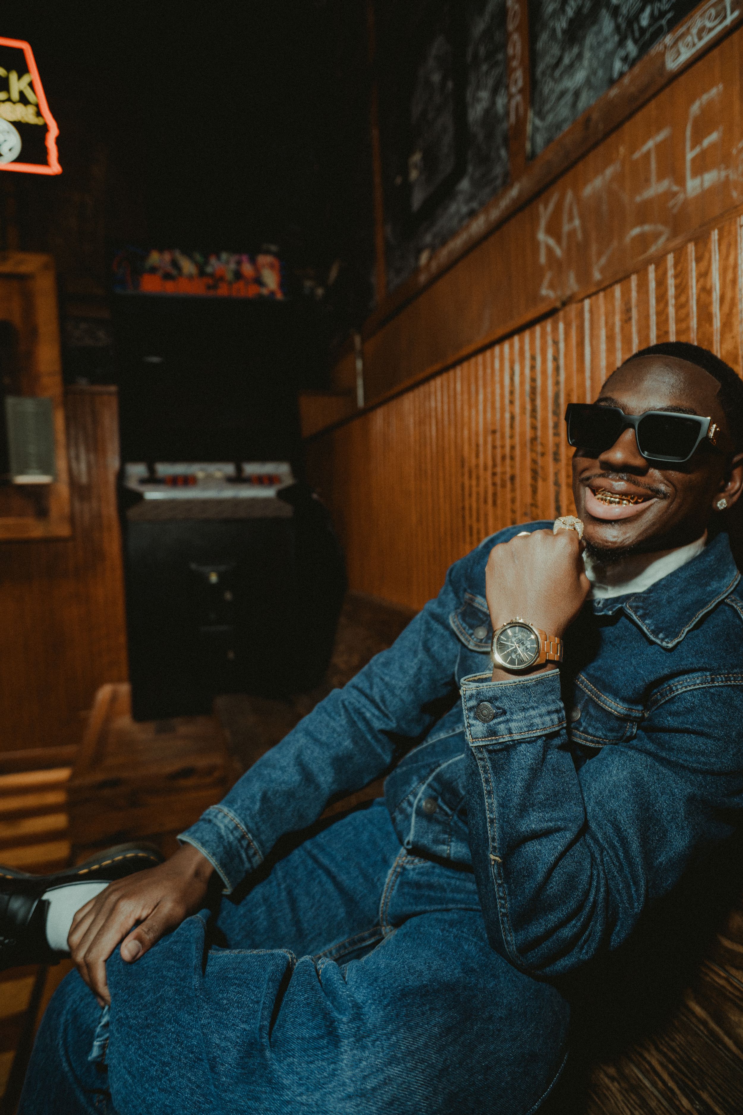 A person in denim clothing smiles while relaxing in a dimly lit room with wooden paneling.