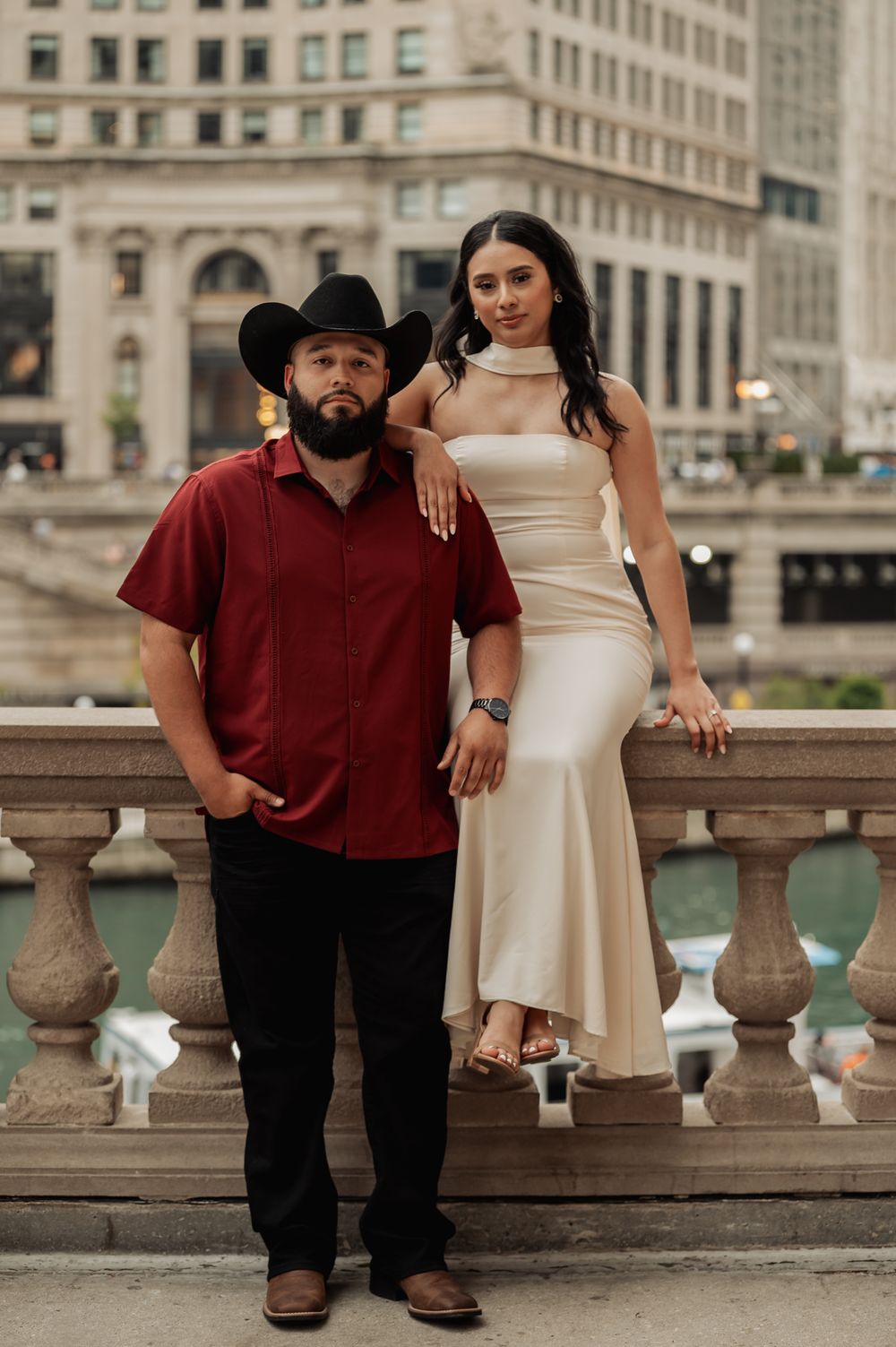 A couple poses together on a stone balcony with urban architecture in the background.