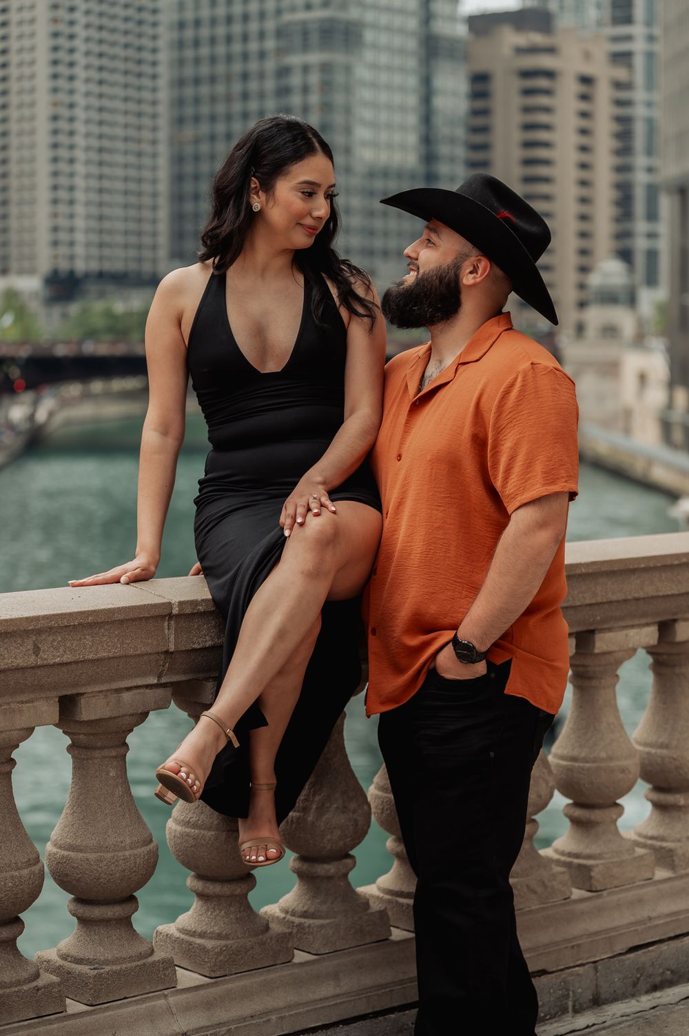 A couple in stylish attire pose together on a stone balcony overlooking a city river with skyscrapers in the background.