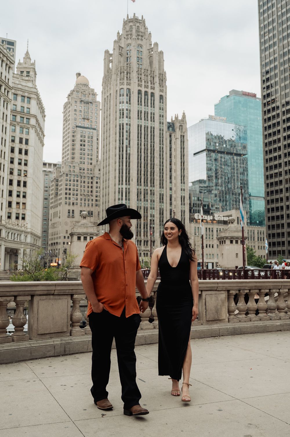 A couple walks along Chicago's riverwalk with the iconic Tribune Tower and skyline behind them.