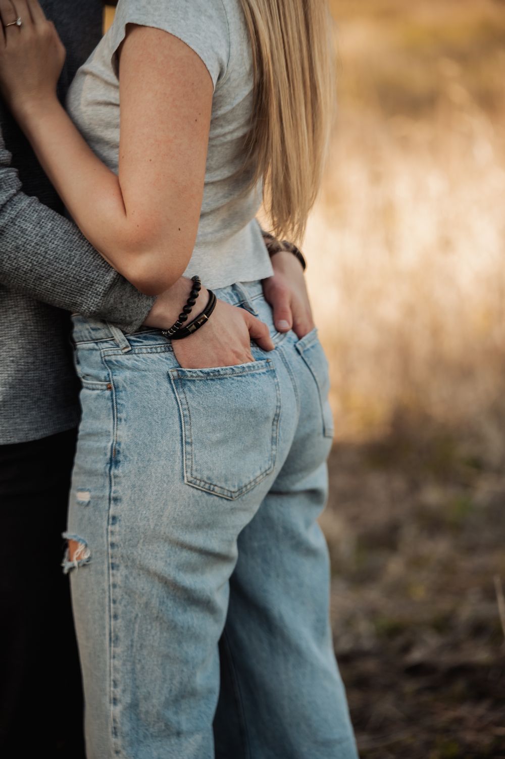 Close-up view of someone wearing light wash denim jeans with hands in back pockets against blurred outdoor background.