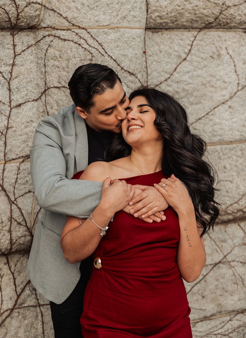 A romantic couple embraces against a textured wall with climbing vines, the woman wearing a burgundy dress.