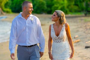 A couple walks hand in hand on the beach at sunset wearing wedding attire.