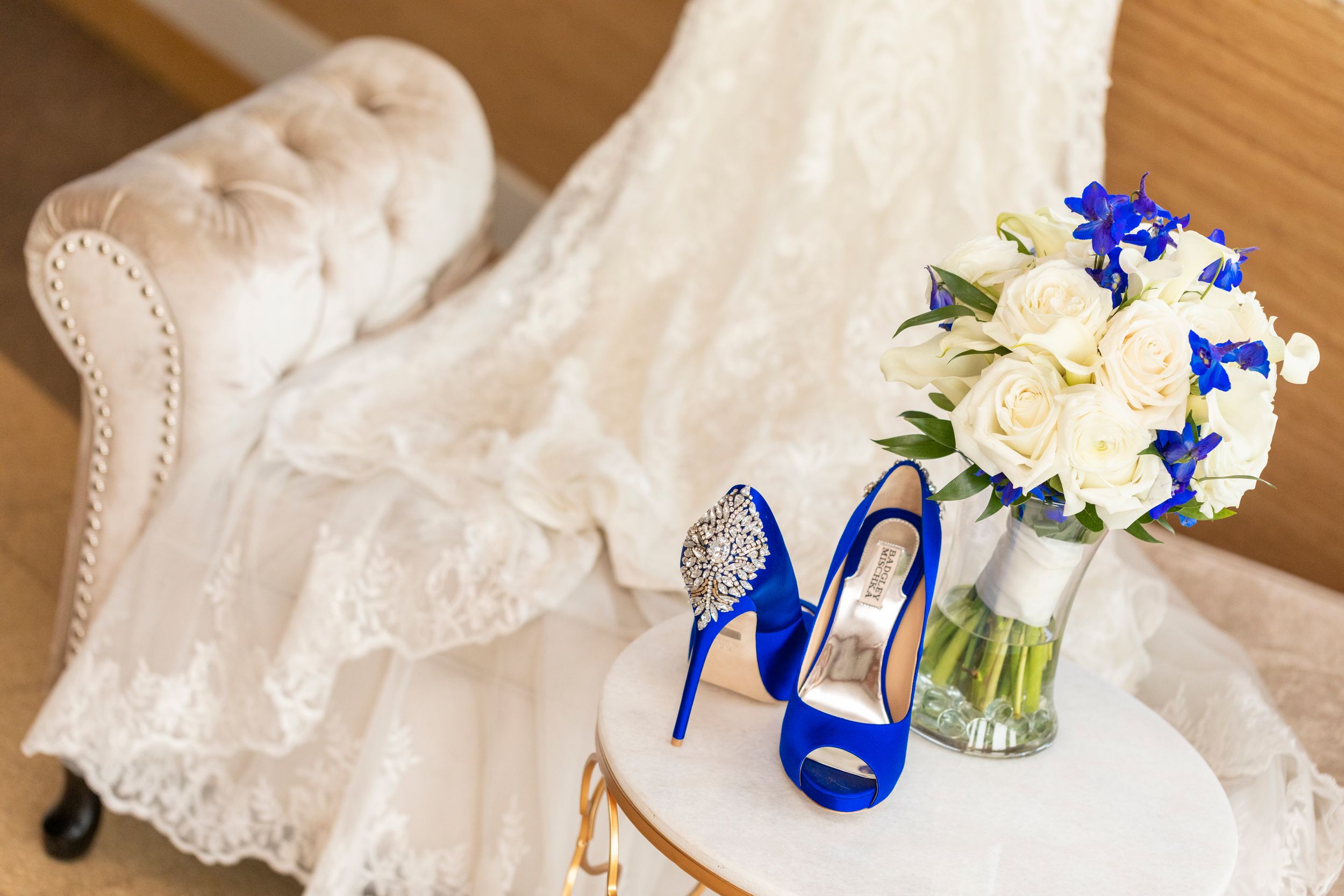 White lace wedding dress with bright blue high heels and floral bouquet arranged on elegant chair.
