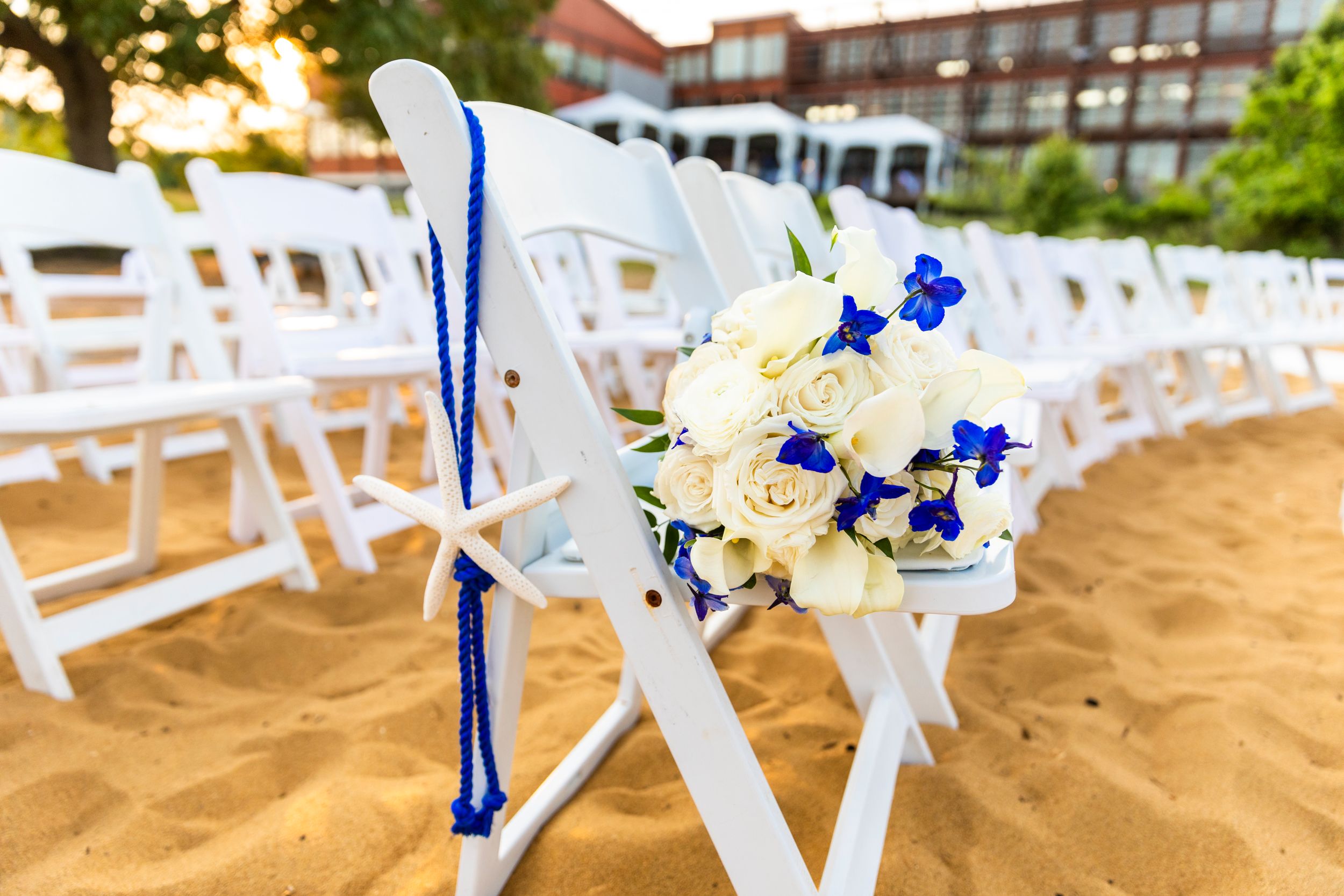 White folding chairs adorned with blue and white floral arrangements on a sandy beach.
