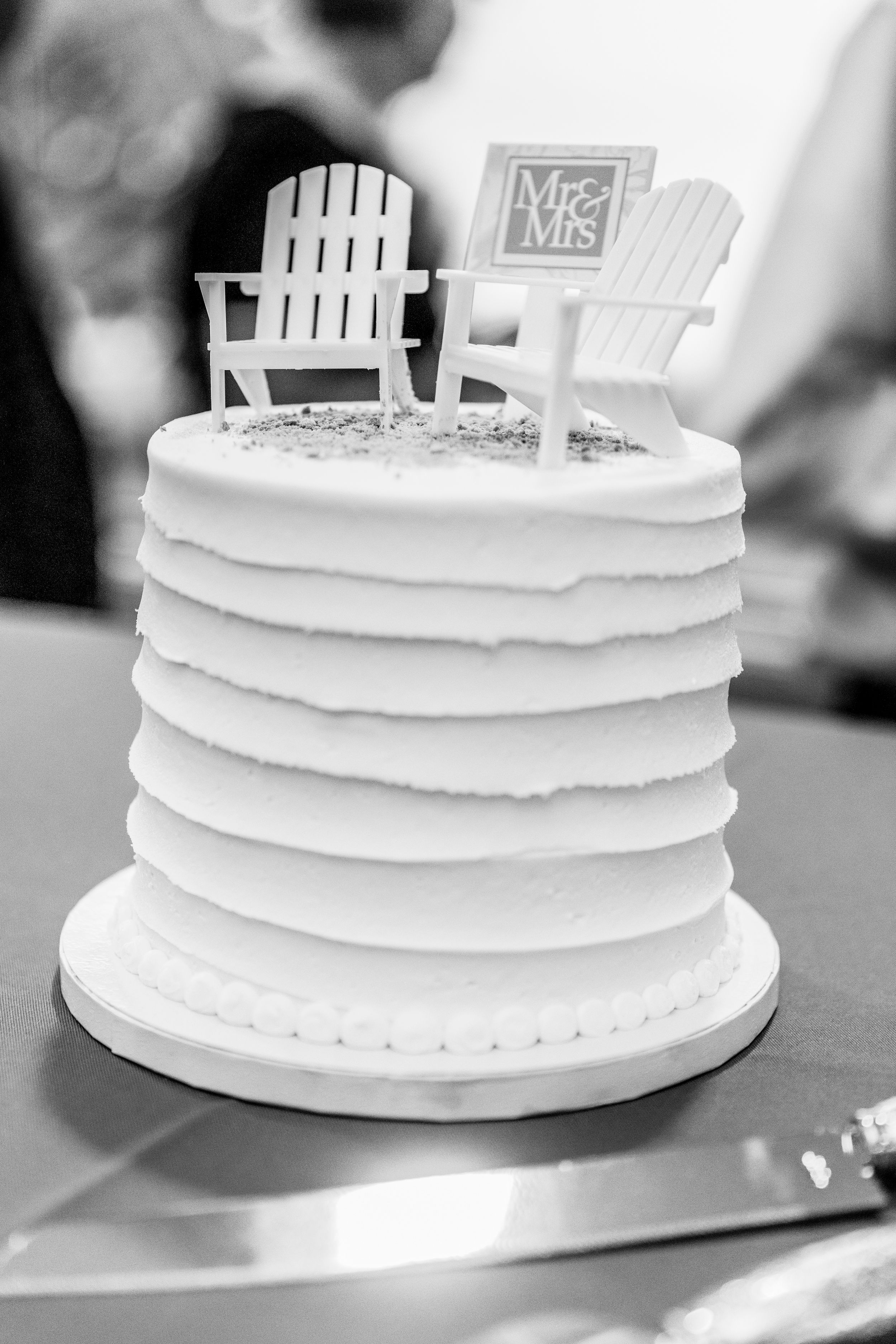 A white wedding cake topped with miniature Adirondack chairs and a Mr & Mrs sign.