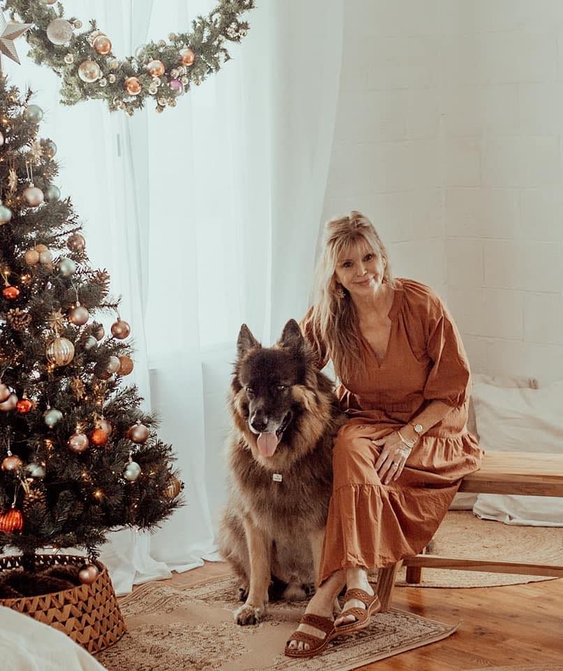 Woman in silk dress sits with German Shepherd by decorated Christmas tree in bright modern room.