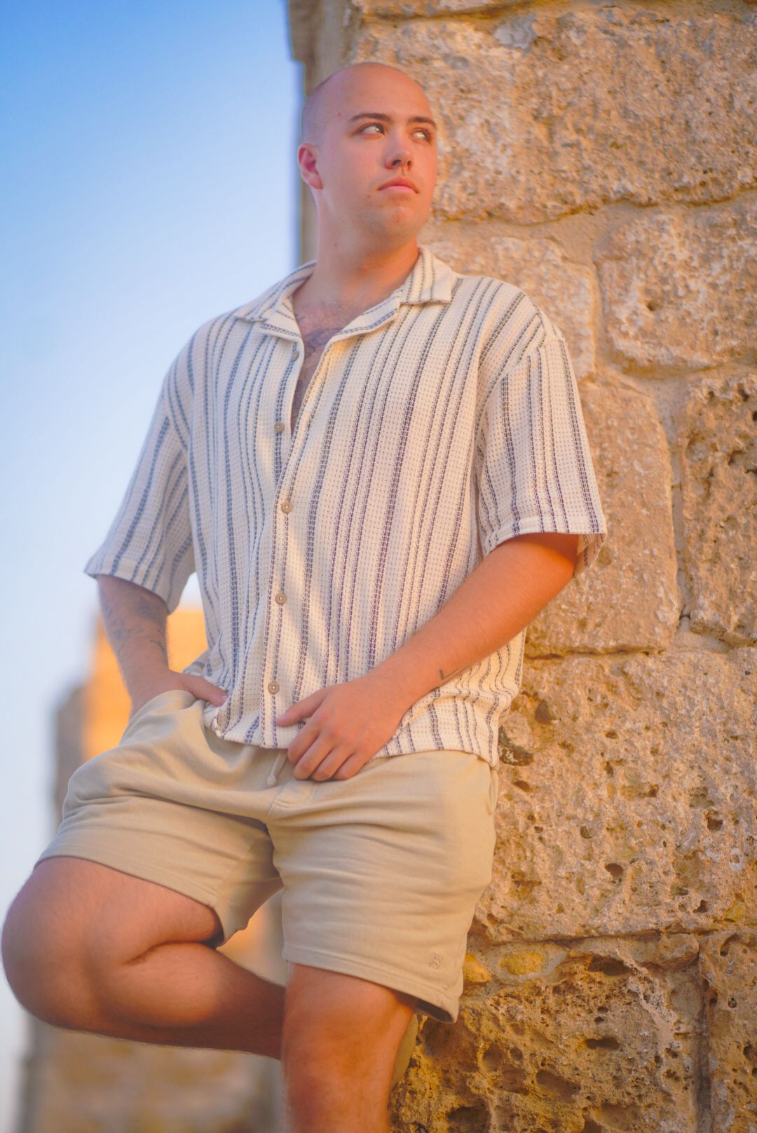 Person in striped shirt and khaki shorts sits casually against a stone wall in natural lighting.