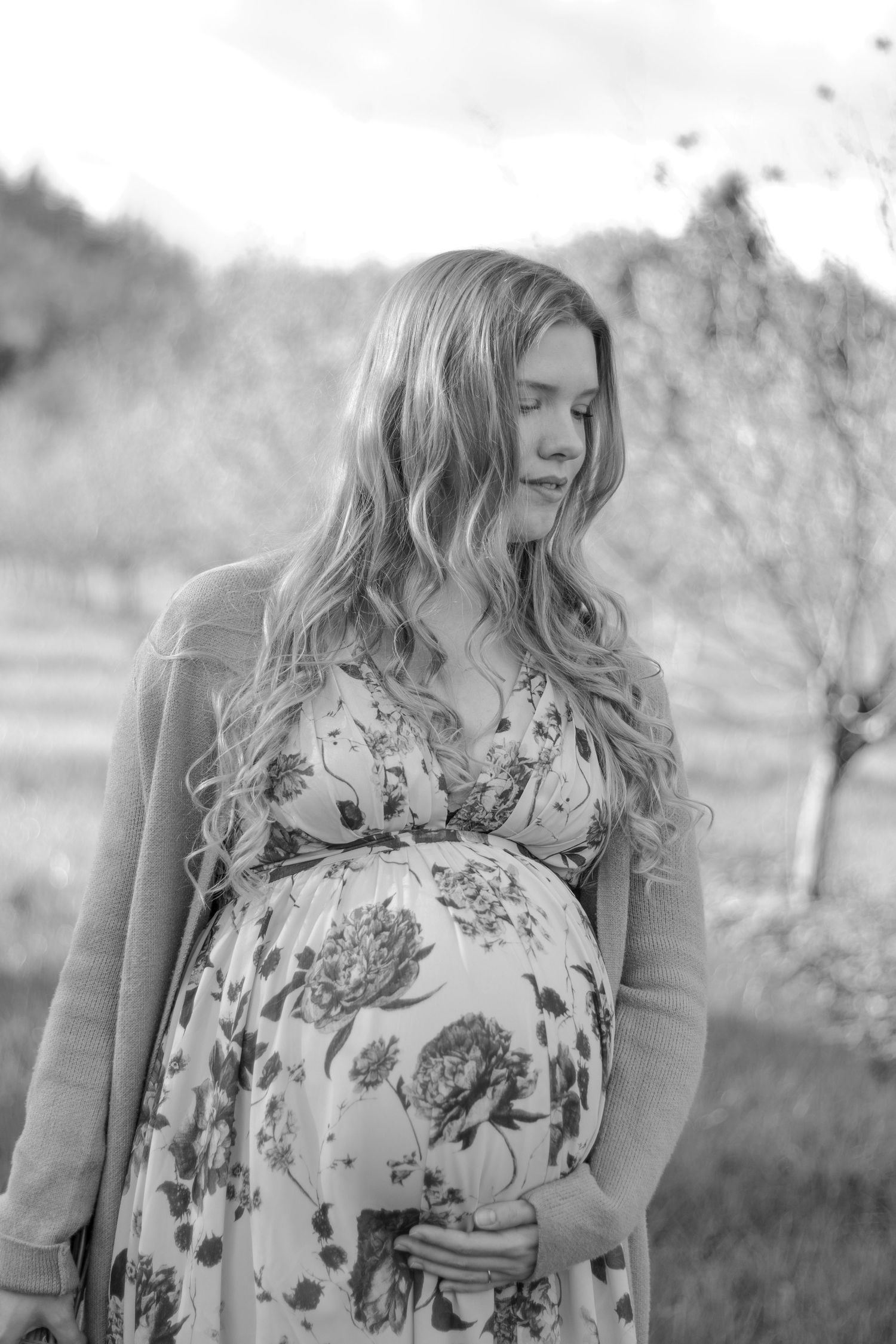Black and white portrait of pregnant woman in floral maxi dress touching belly in garden setting.