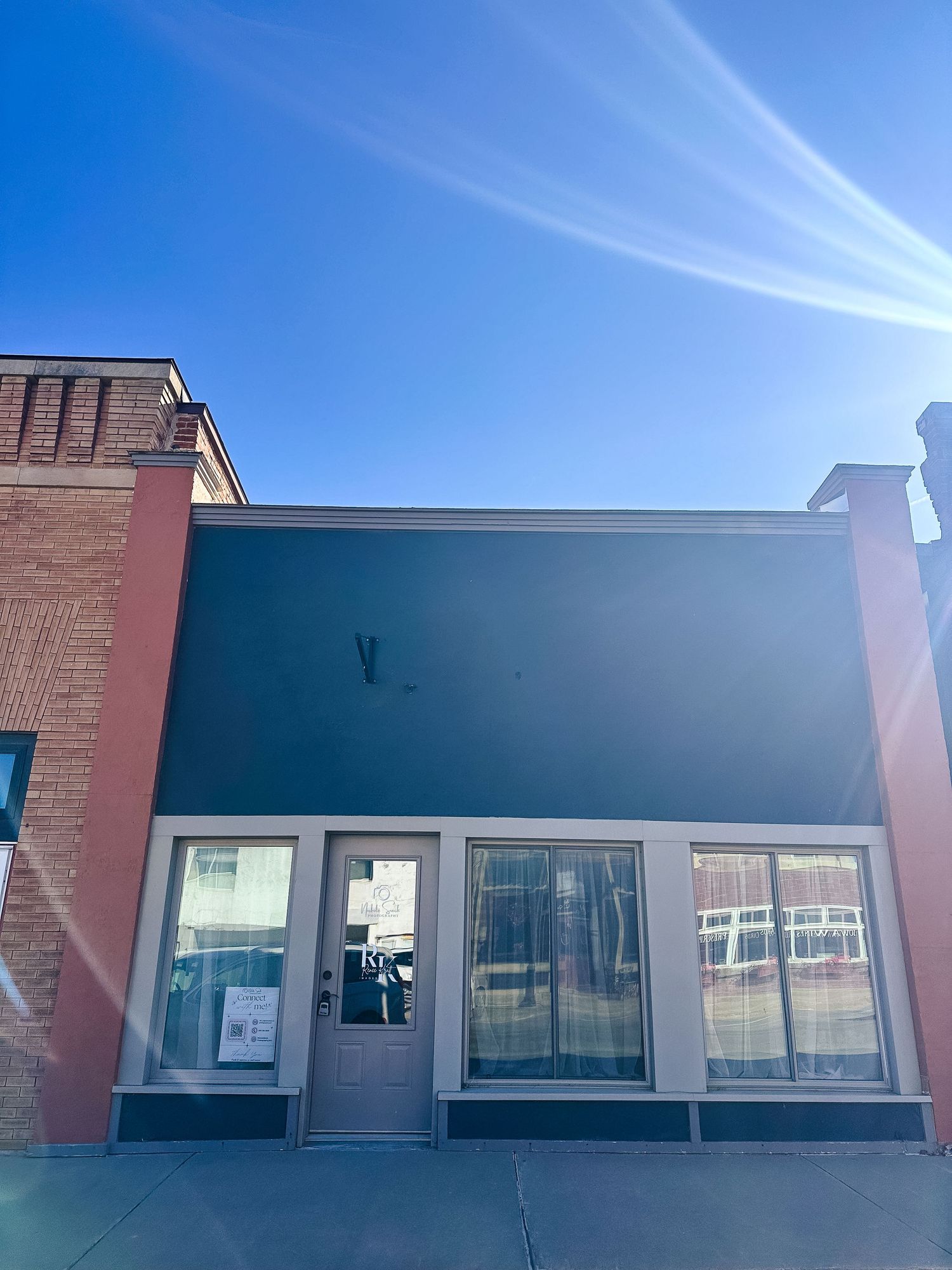 Storefront of a small brick building with dark blue facade and glass windows under bright blue sky with wispy clouds.