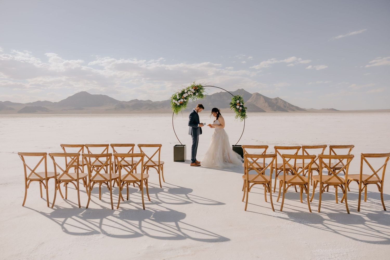 Minimalist outdoor wedding ceremony on white salt flats with circular floral arch and wooden chairs.