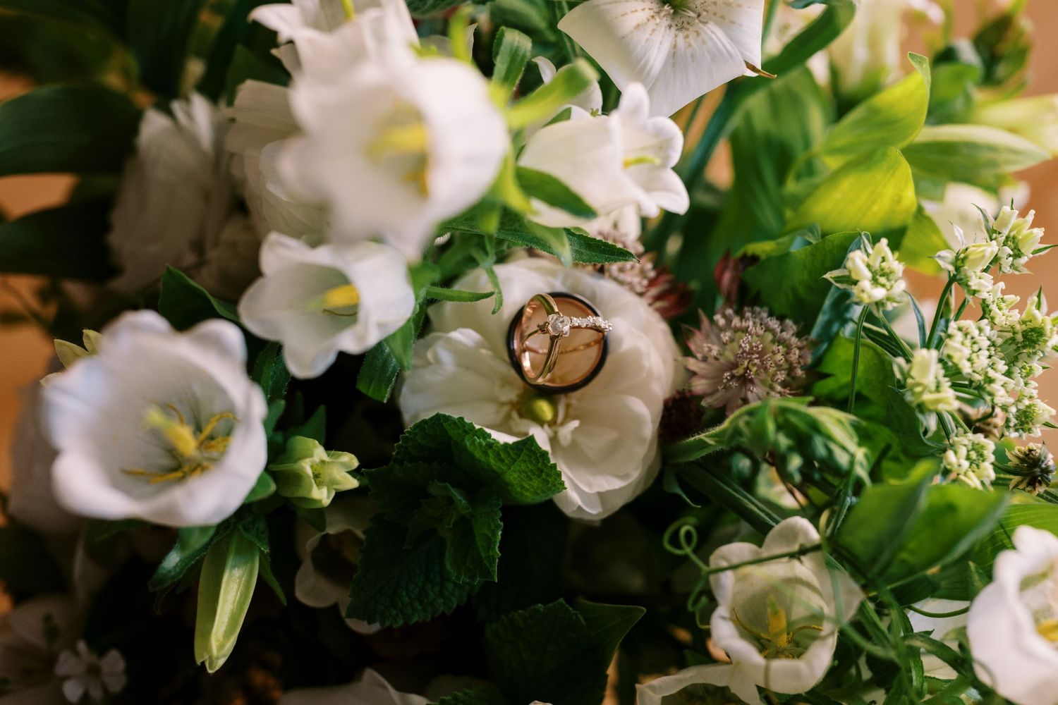 Gold wedding rings nestled among white bell-shaped flowers and green foliage in a romantic floral arrangement.