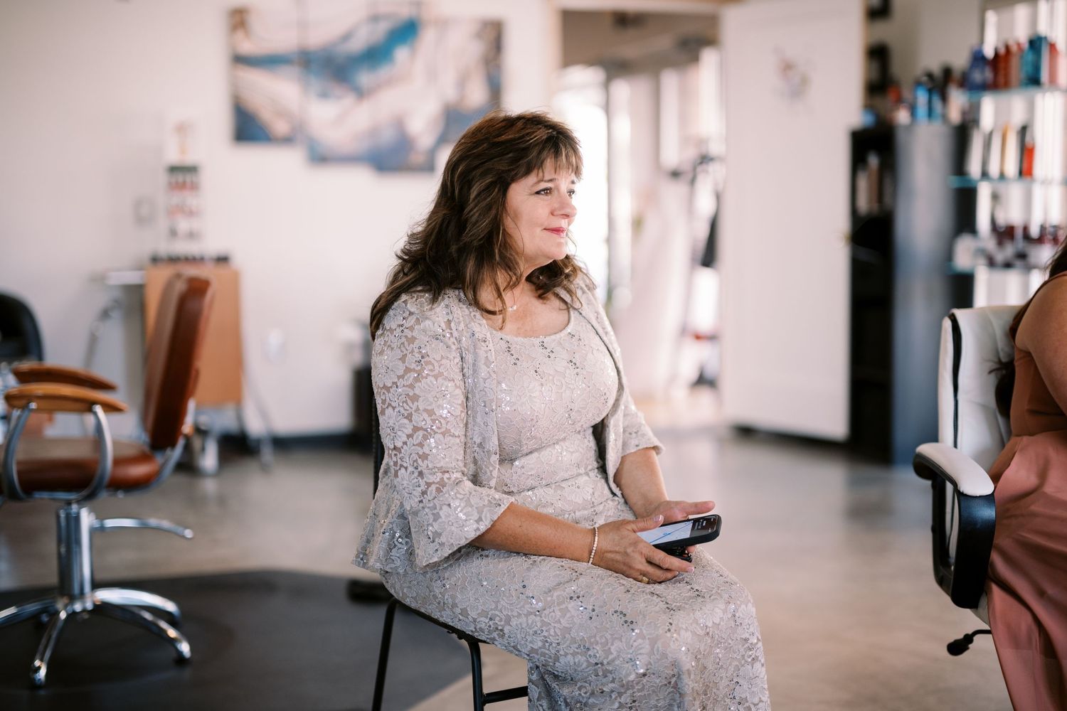 Someone sits in a salon chair wearing a light gray lace dress while looking at their phone.