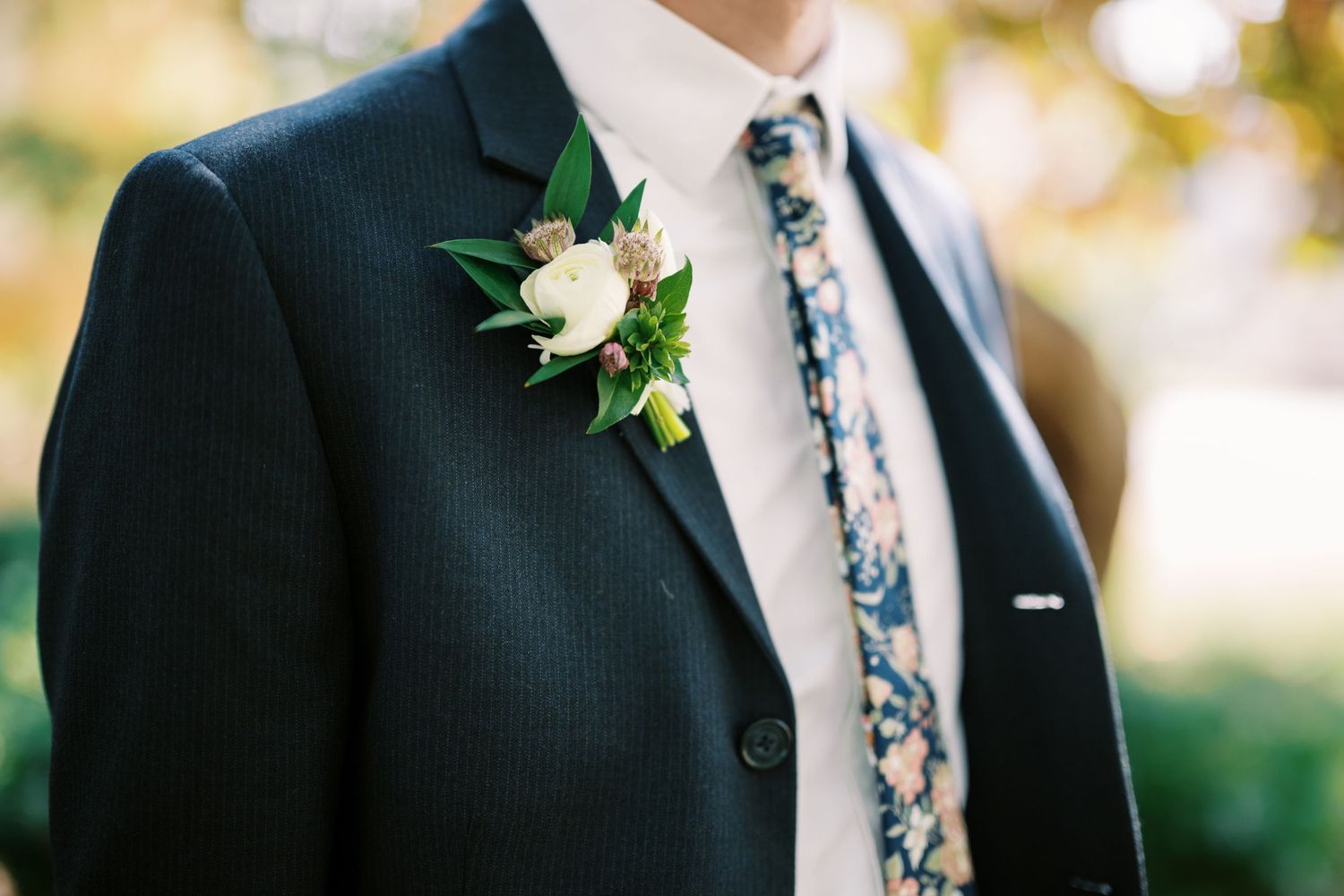 Close-up of a black suit jacket with white boutonniere and floral patterned tie against blurred outdoor background.