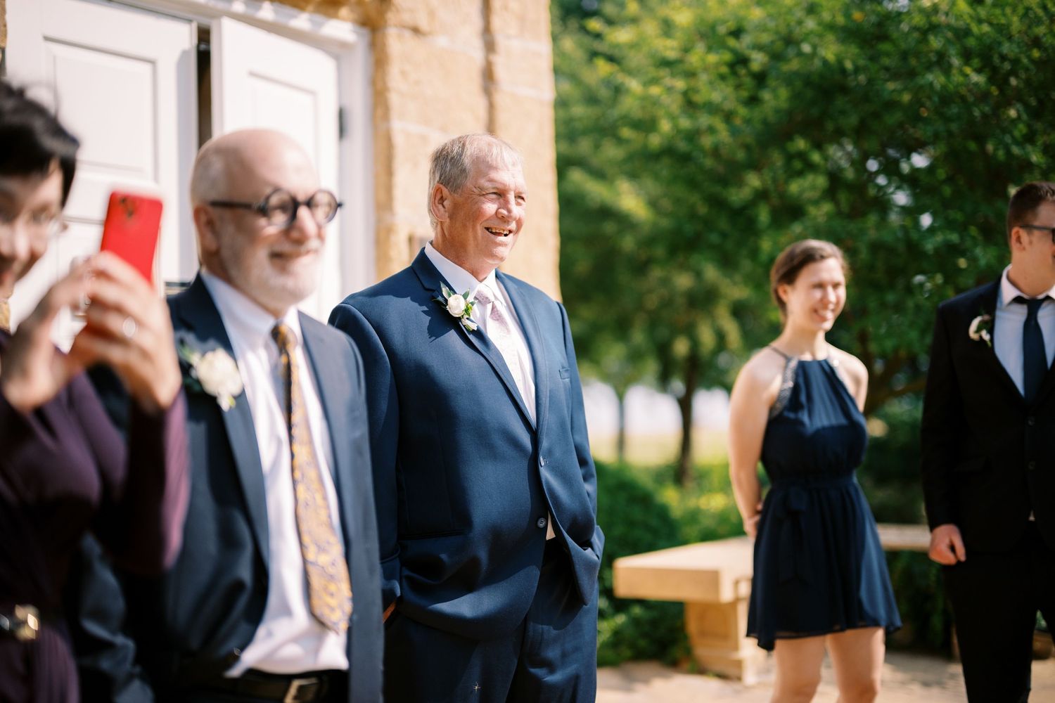 Well-dressed guests gather outside at an elegant wedding celebration in a sunny garden setting.