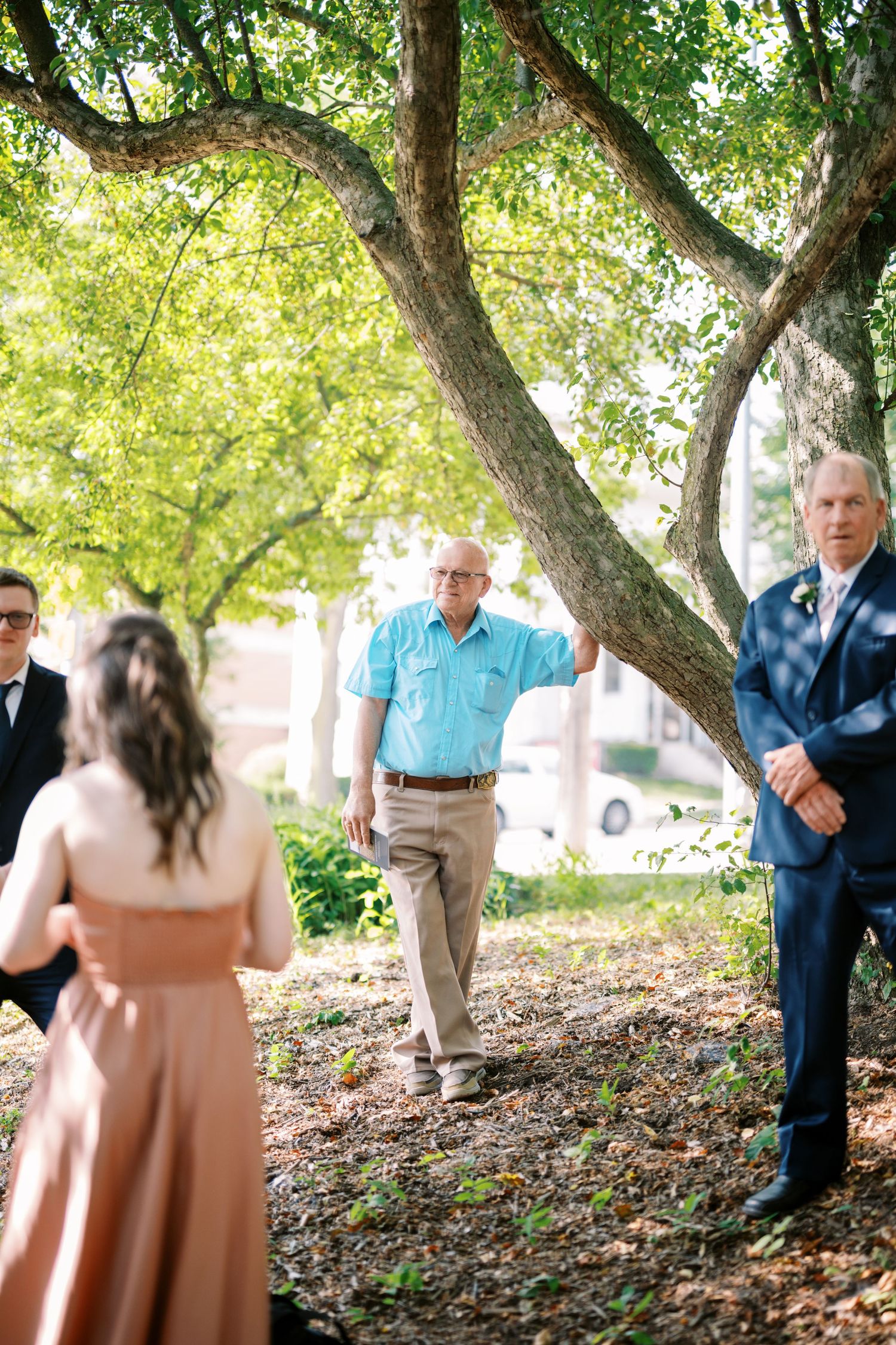 A small outdoor gathering under a large shady tree on a sunny day, with people standing on fallen leaves.