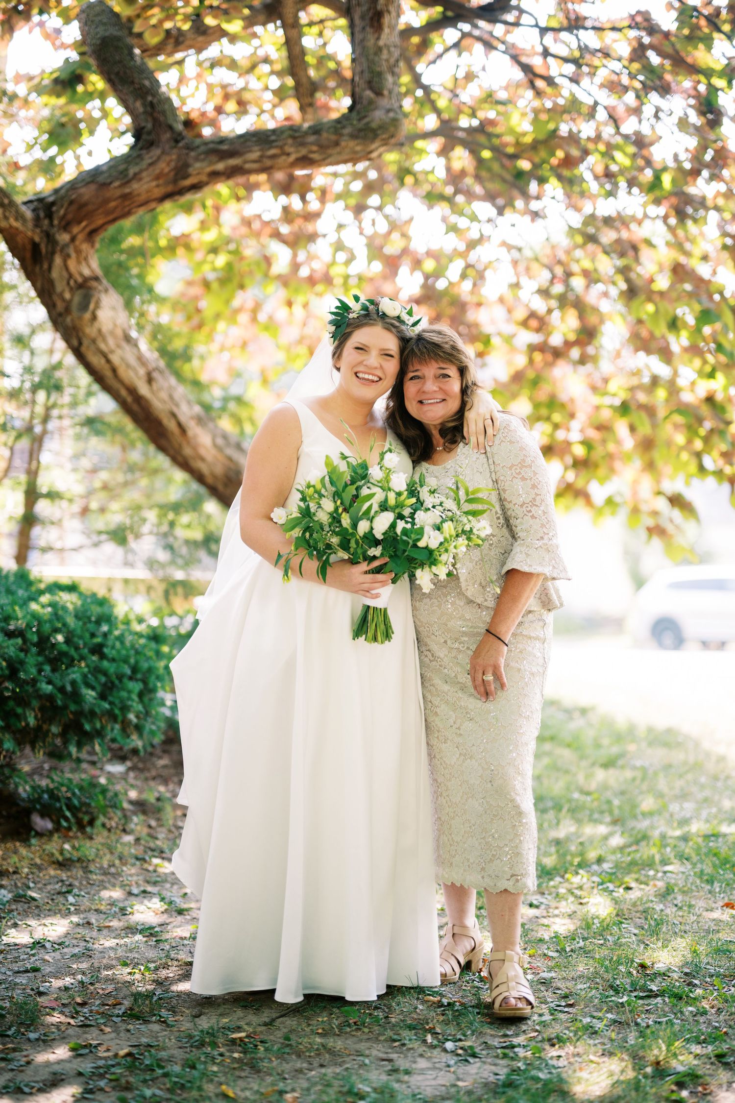 A wedding photo outdoors under a tree in autumn with greenery and white floral bouquet.