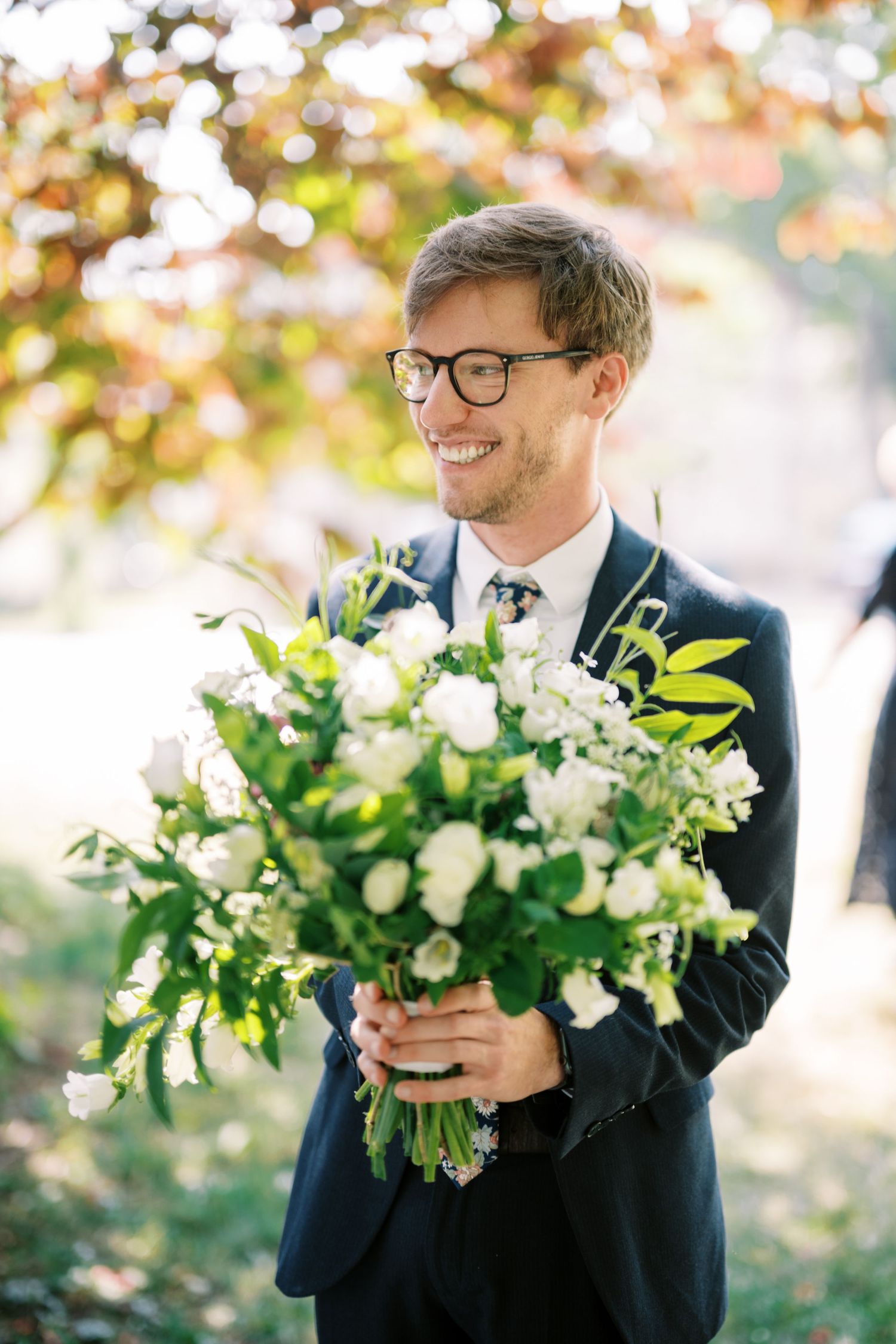 Joyful groom in navy suit and glasses holds beautiful white floral bouquet during outdoor autumn wedding ceremony.