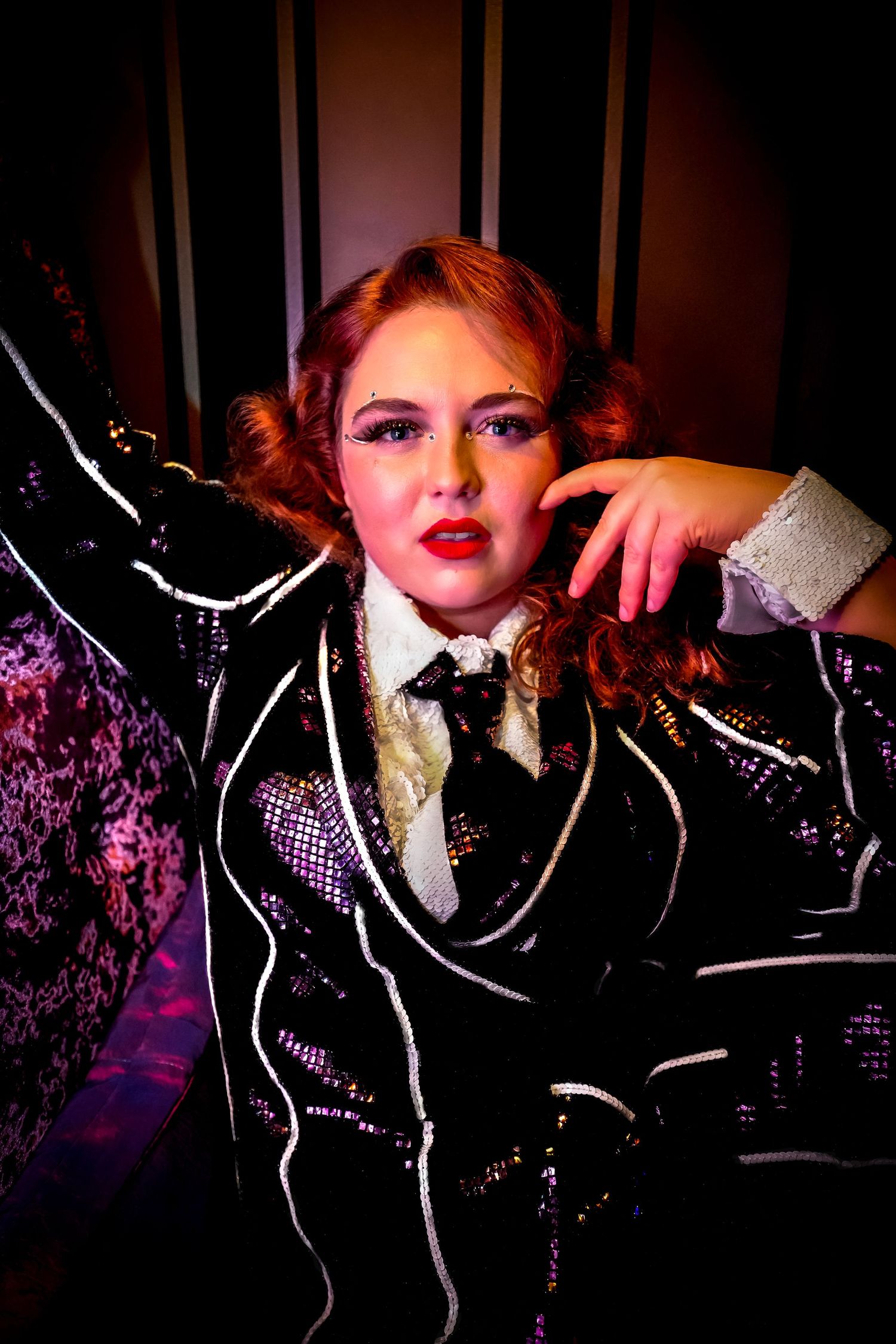 Glamorous vintage-inspired portrait featuring dramatic lighting, bold red lips, and a sparkly black suit with bow tie.