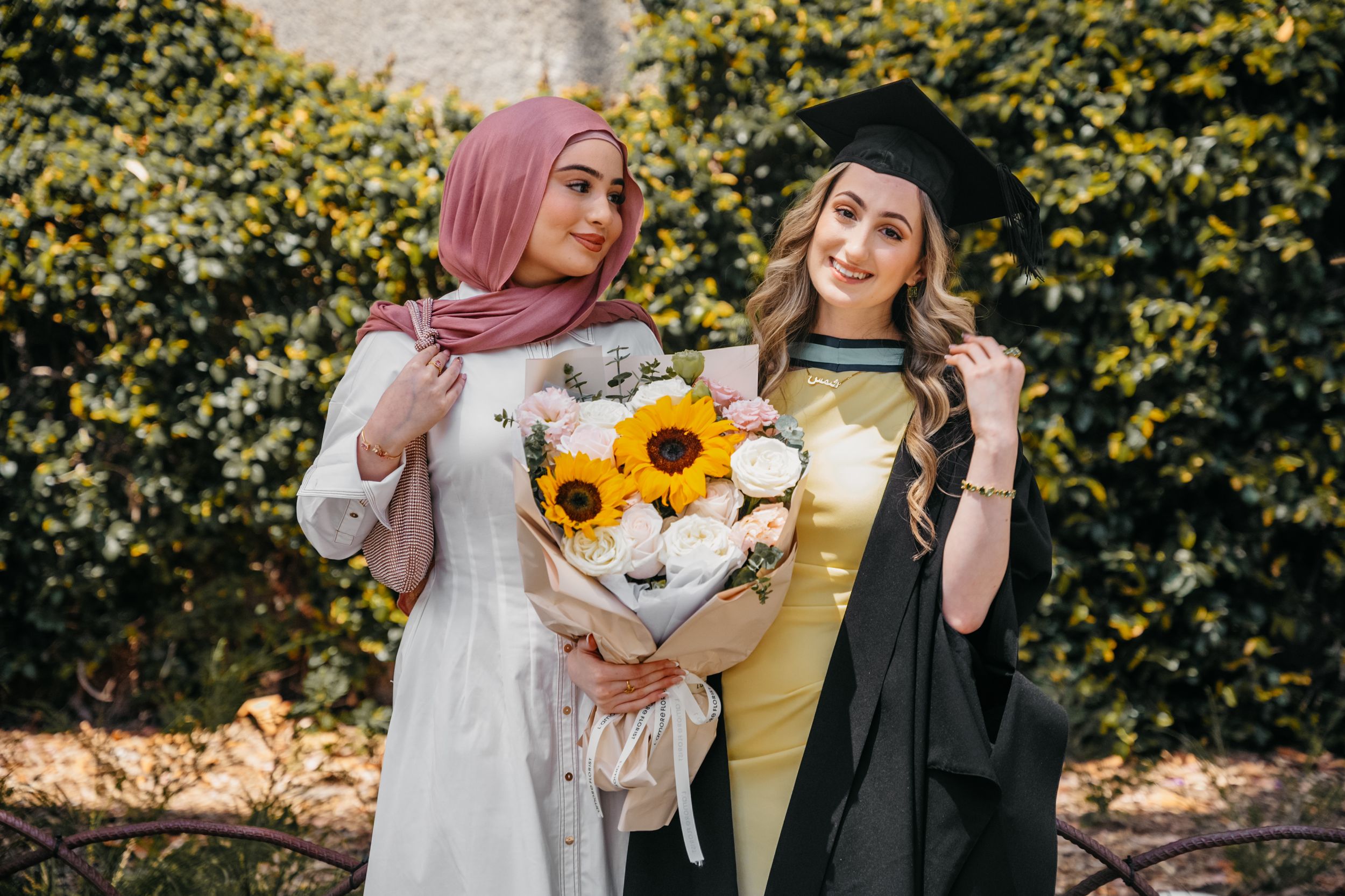 Two graduates in academic regalia holding sunflowers pose together outdoors.