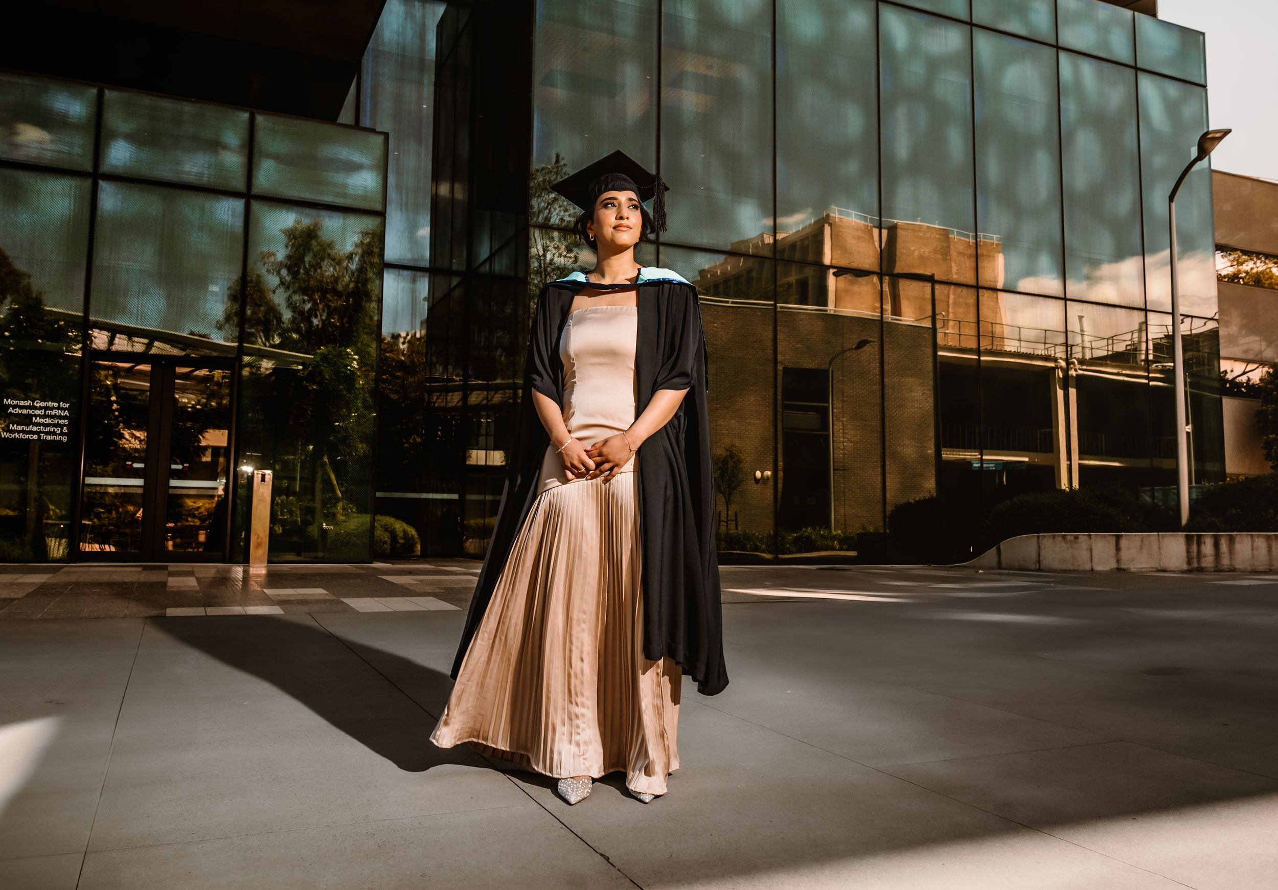 Graduate in academic dress poses in front of modern glass building during golden hour.
