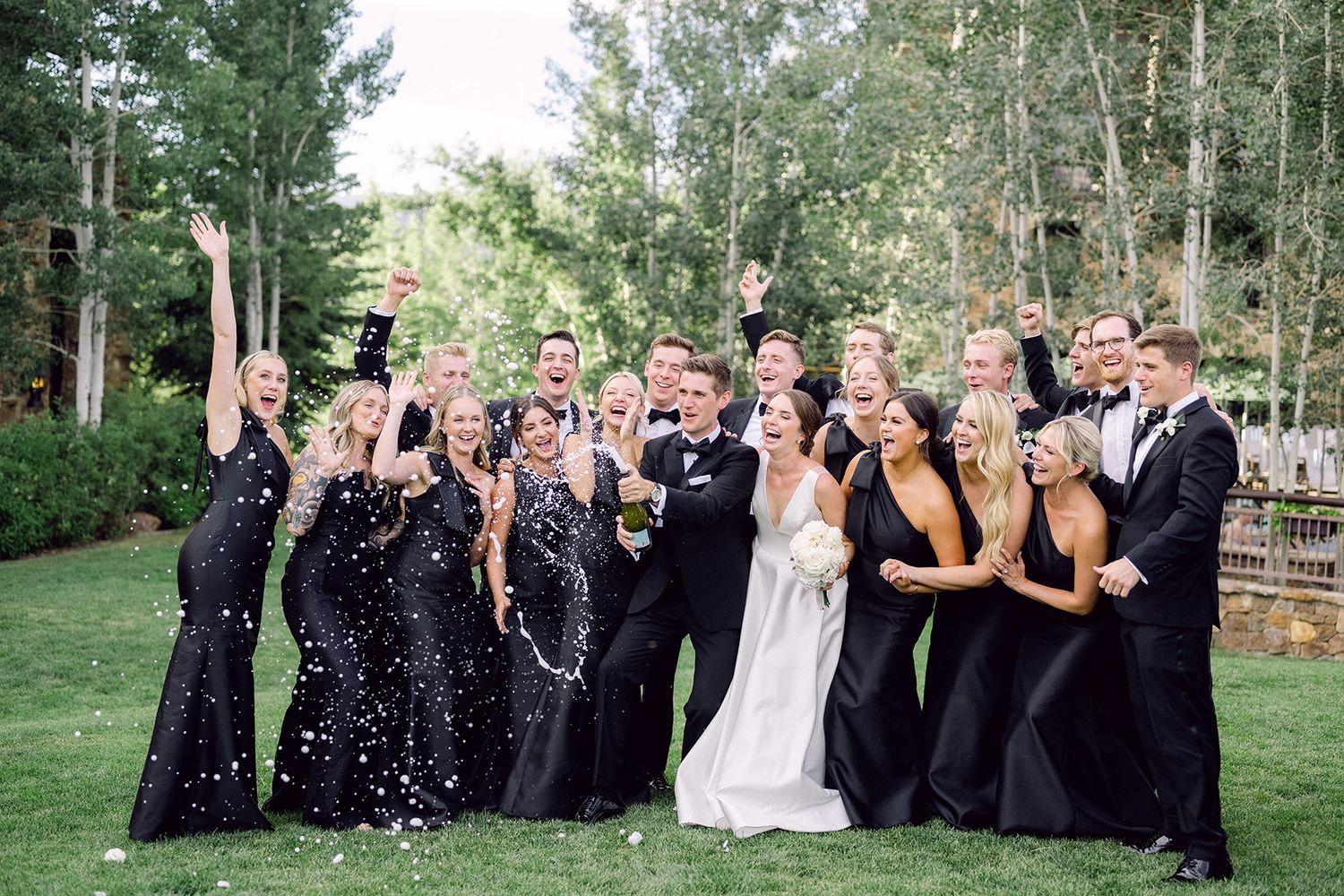 Wedding party celebrating with champagne in formal black attire against garden background.
