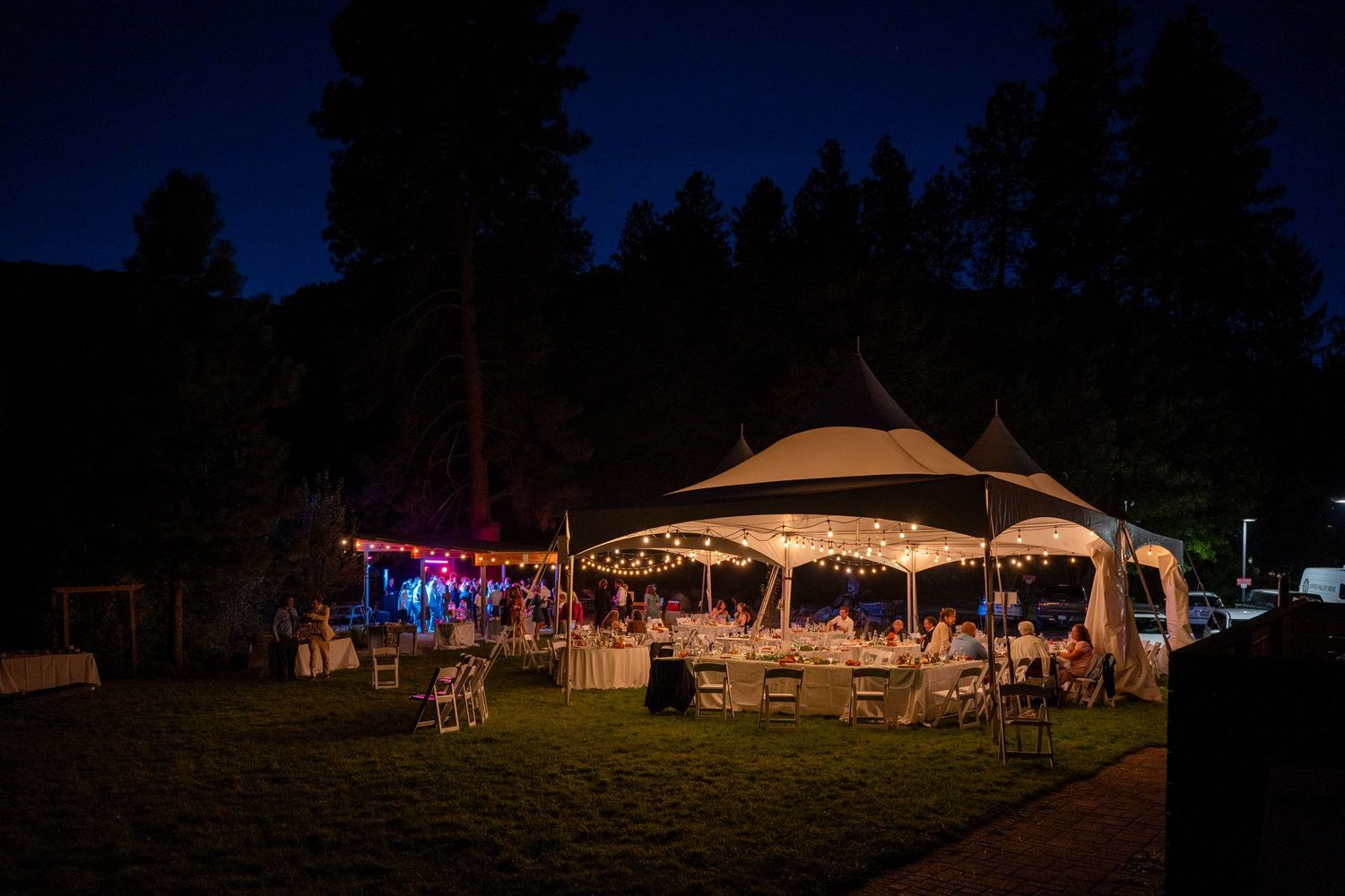 An outdoor wedding reception tent illuminated with string lights at dusk.